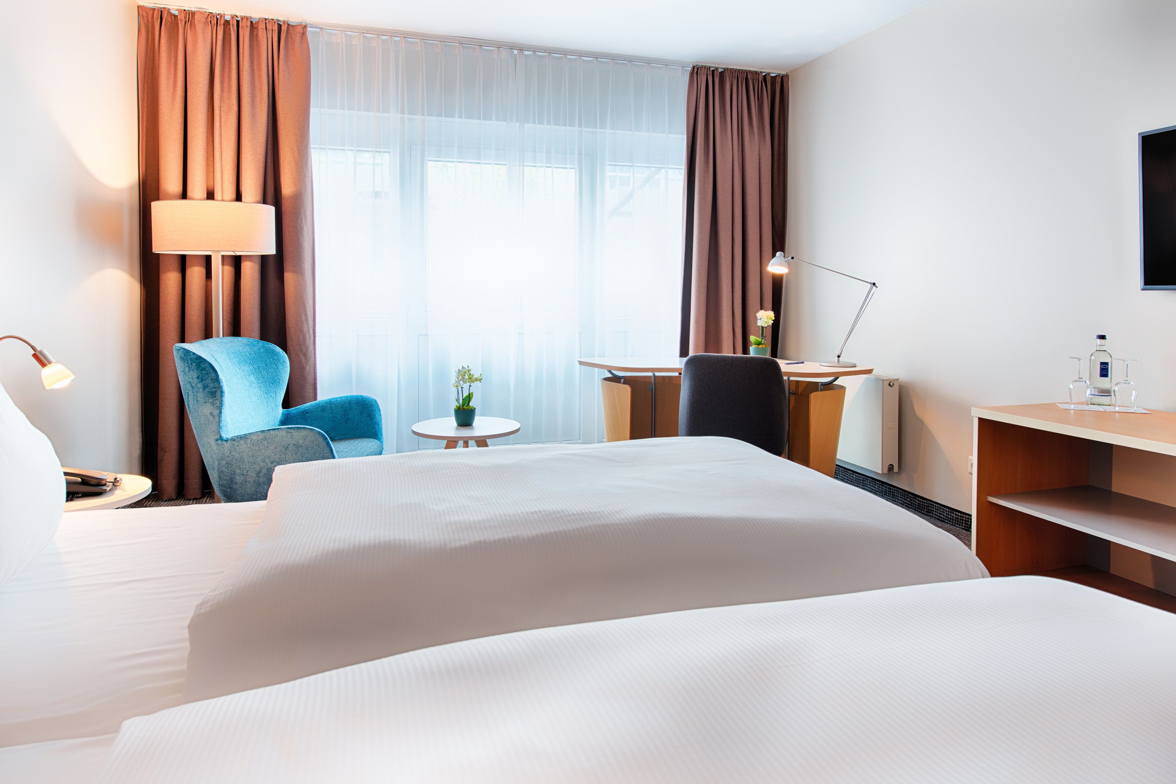 Une chambre d’hôtel avec un grand lit, un bureau avec une chaise, une petite table avec une plante et une lampe.