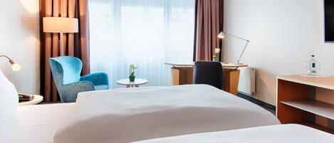 A hotel room with a large bed, a desk with a chair, a small table with a plant, and a lamp.
