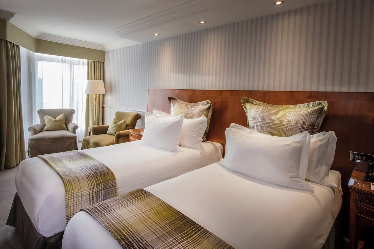 A hotel room with two beds, a wooden headboard, a lamp, and a chair.