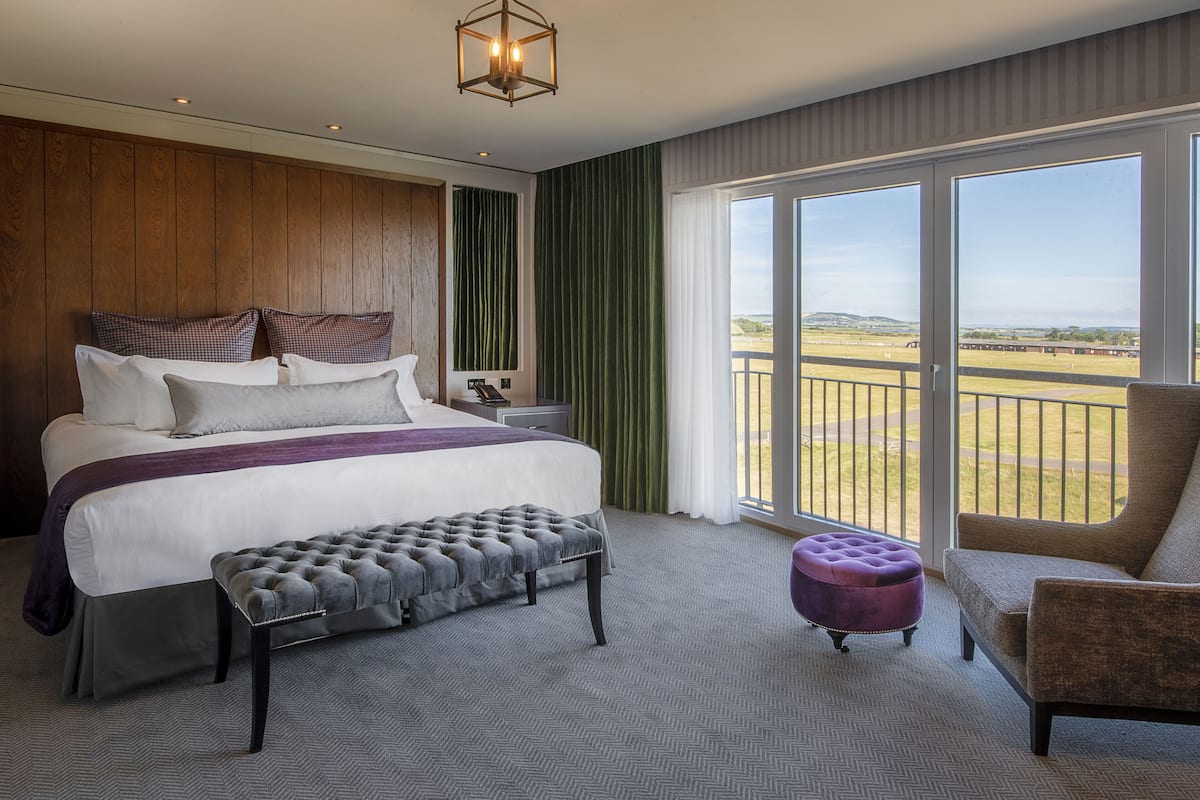 A bedroom with a large bed, a bench, a chair, a nightstand, and a view of the outdoors.