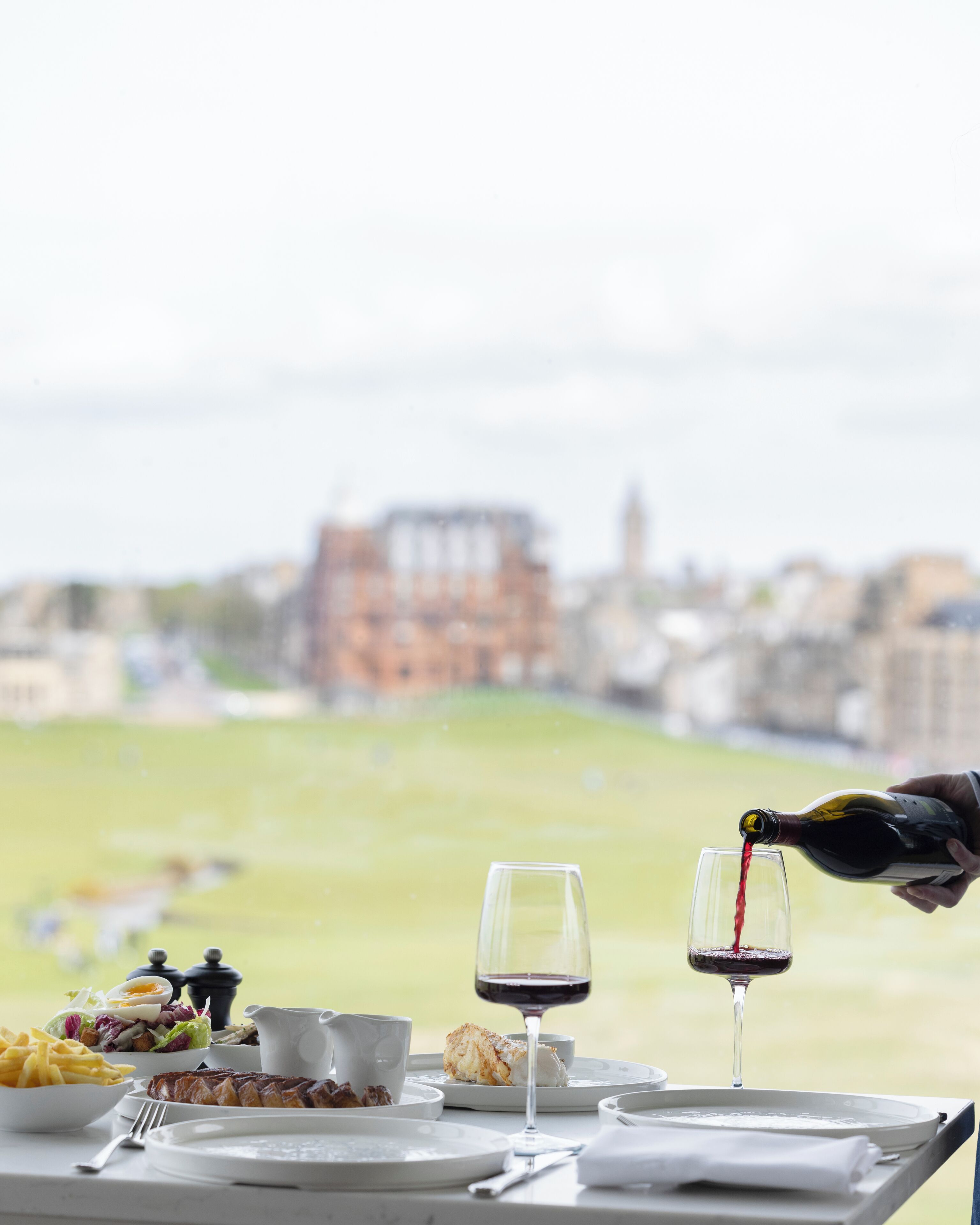A table set with a meal, two glasses of red wine, and a bottle being poured.