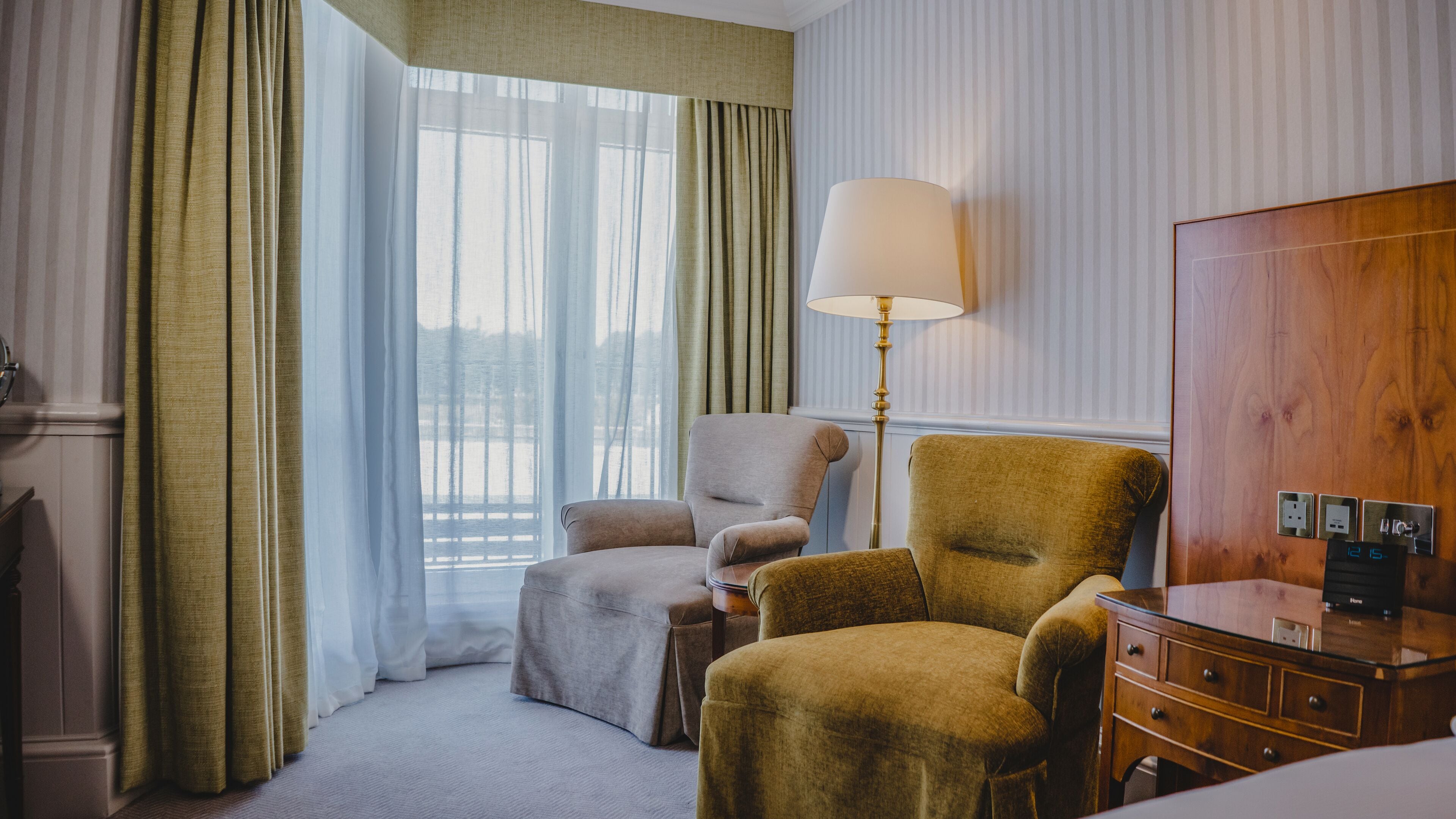 A hotel room with a large window, a lamp, a wooden wardrobe, and a desk with a computer.