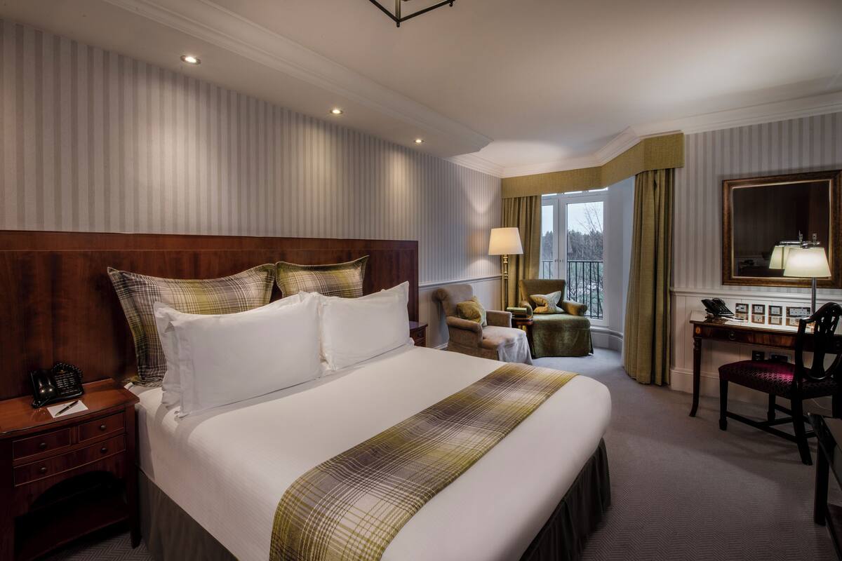 A hotel room with a large bed, a desk with a chair, a window with curtains, and a mirror on the wall.