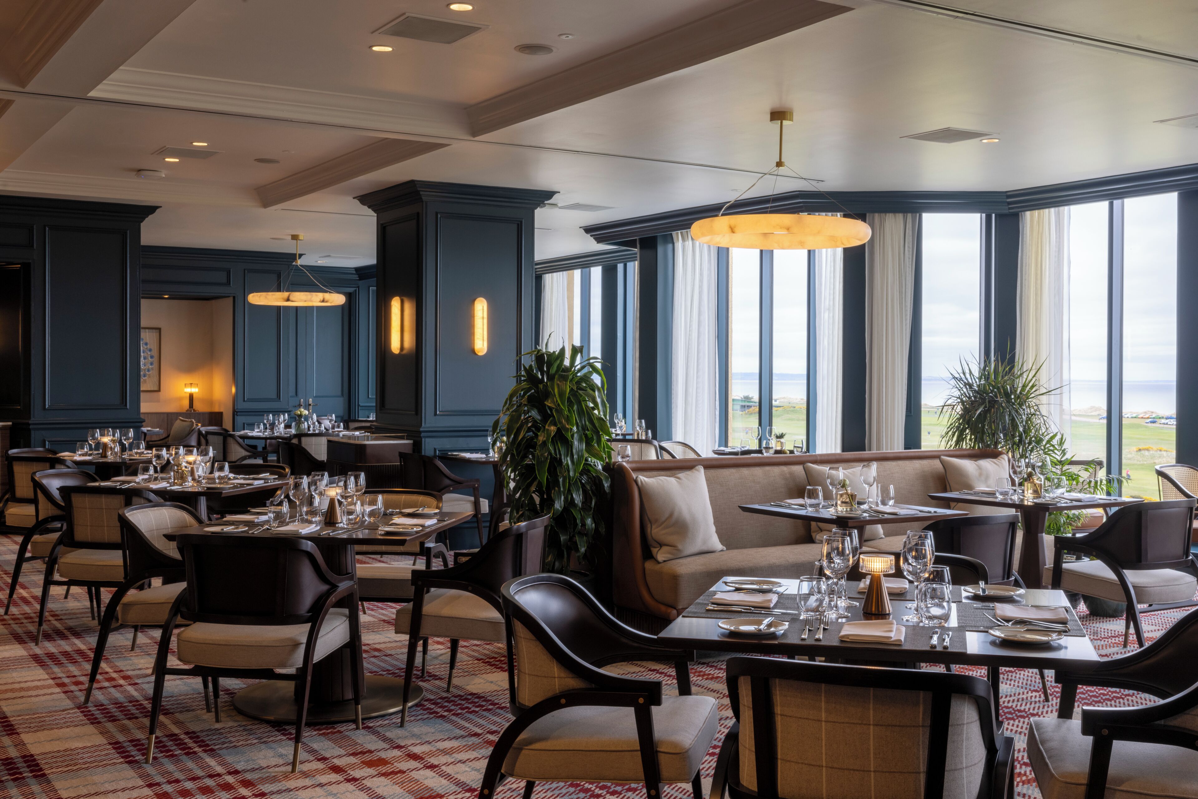 A restaurant with tables set for dining, large windows offering a view, and a high ceiling with recessed lighting.