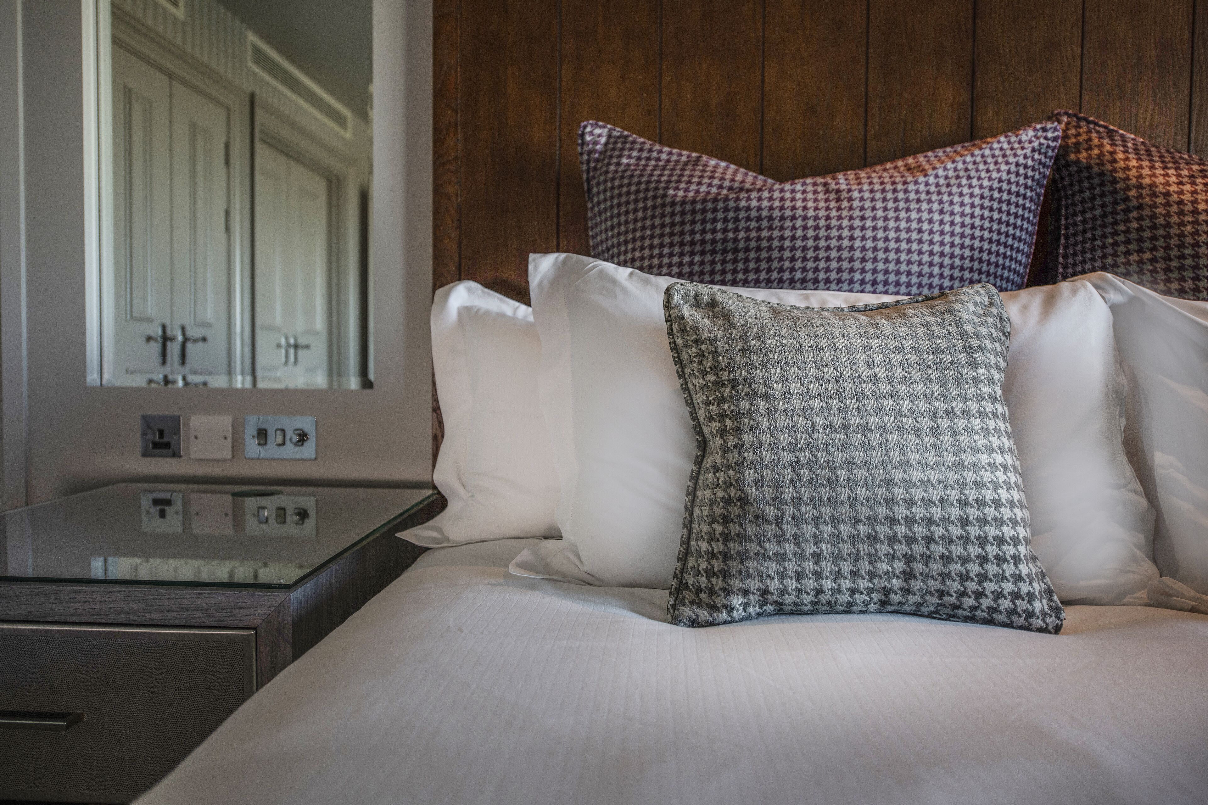 Uma cama bem arrumada com almofadas brancas e estampadas, cabeceira de madeira e mesa de cabeceira com espelho e tomadas.