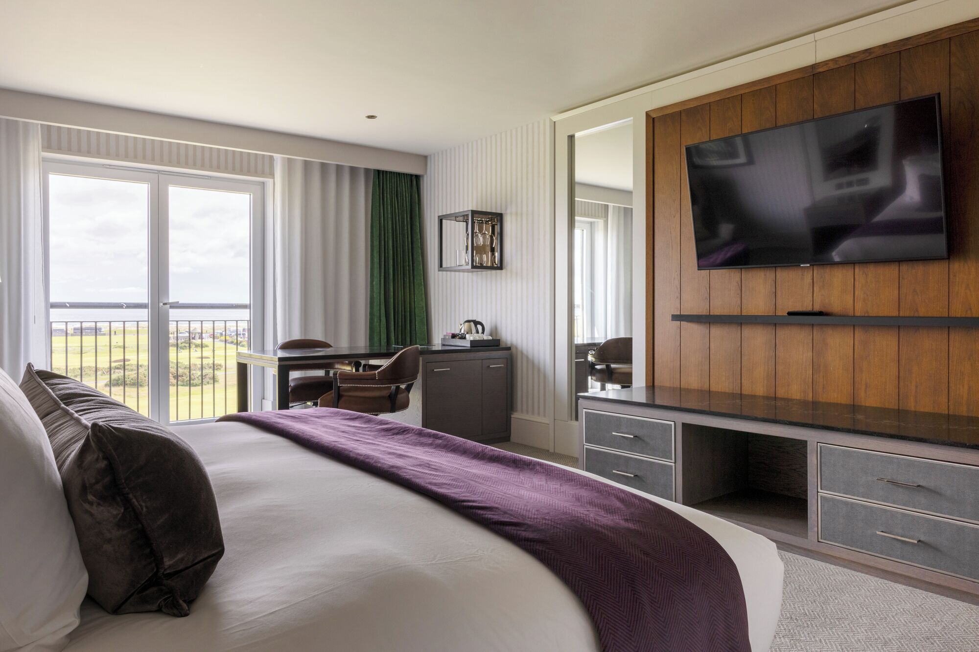 A hotel room with a large bed, a desk with a chair, a TV mounted on the wall, and a view of the outdoors through the window.