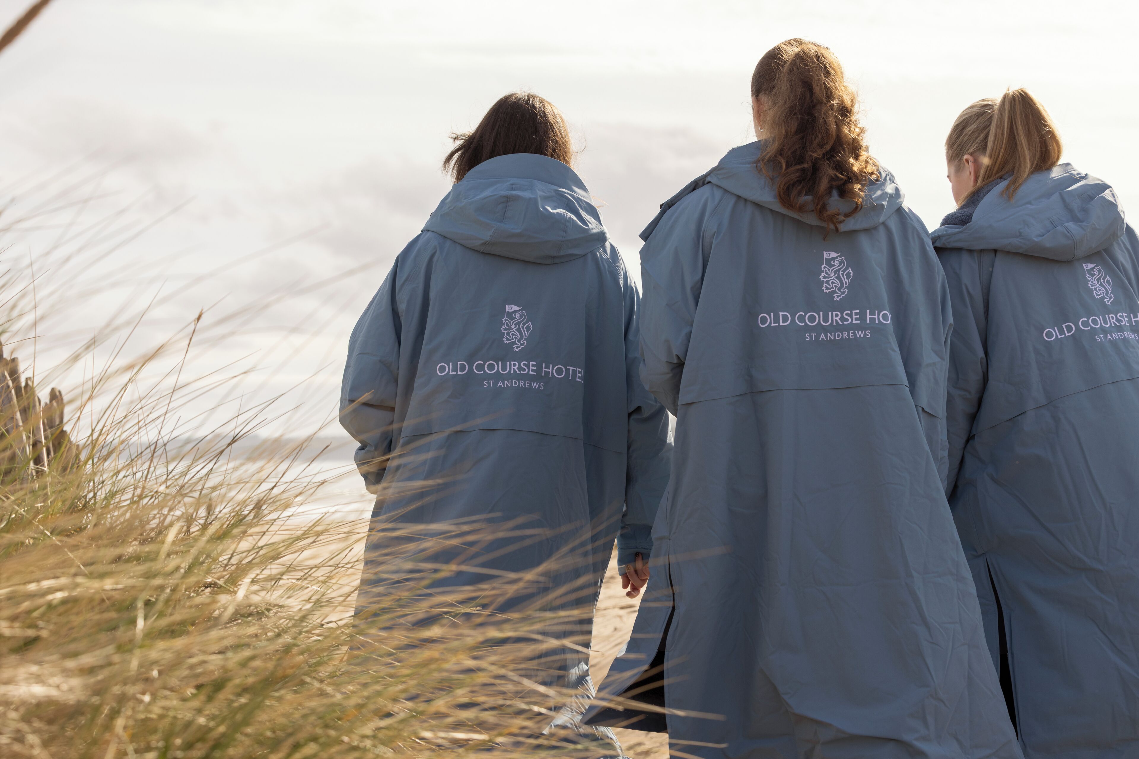 Three individuals wearing jackets with 'Old Course Hotel St Andrews' text, standing on a grassy area with a view of the sea.