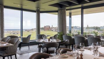 Un restaurant amb taules preparades per dinar, grans finestres que ofereixen vista a un paisatge urbà i un camp de golf.