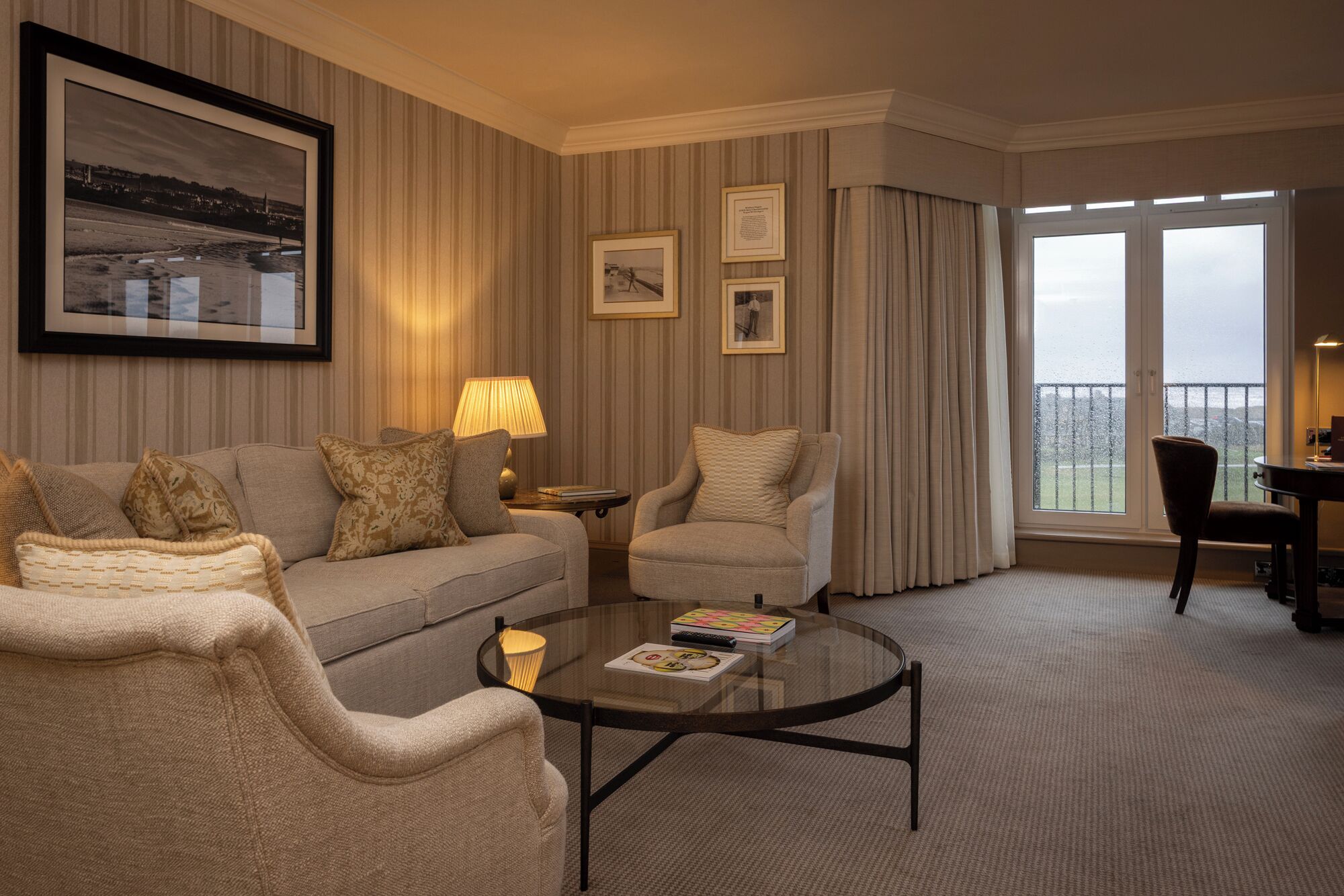 A hotel room with a sofa, armchair, and a glass coffee table with magazines. There are framed pictures on the wall and a view of the outdoors through the window.