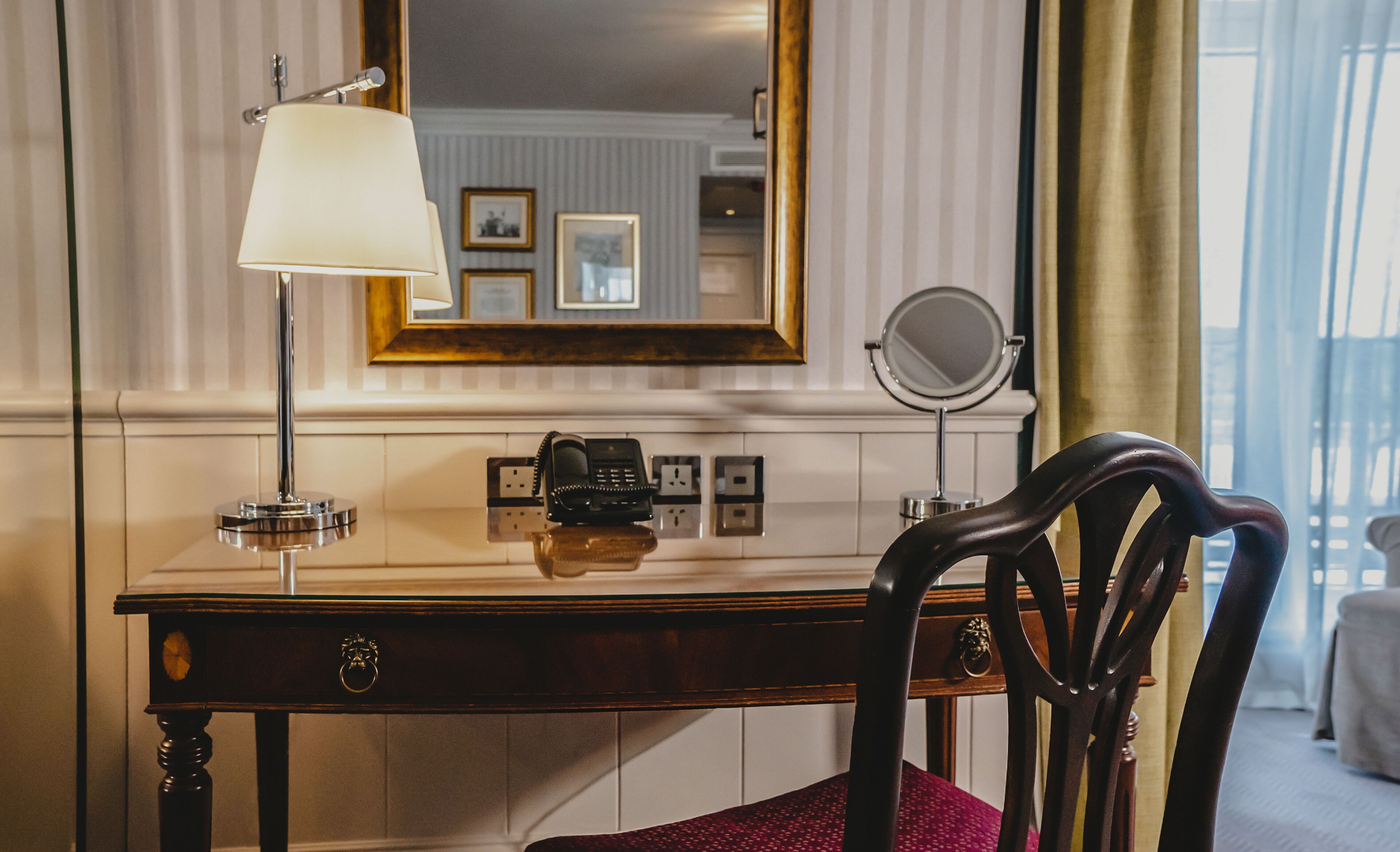 Habitación de hotel con un escritorio de madera, una lámpara, un teléfono, un espejo y una ventana con cortinas.