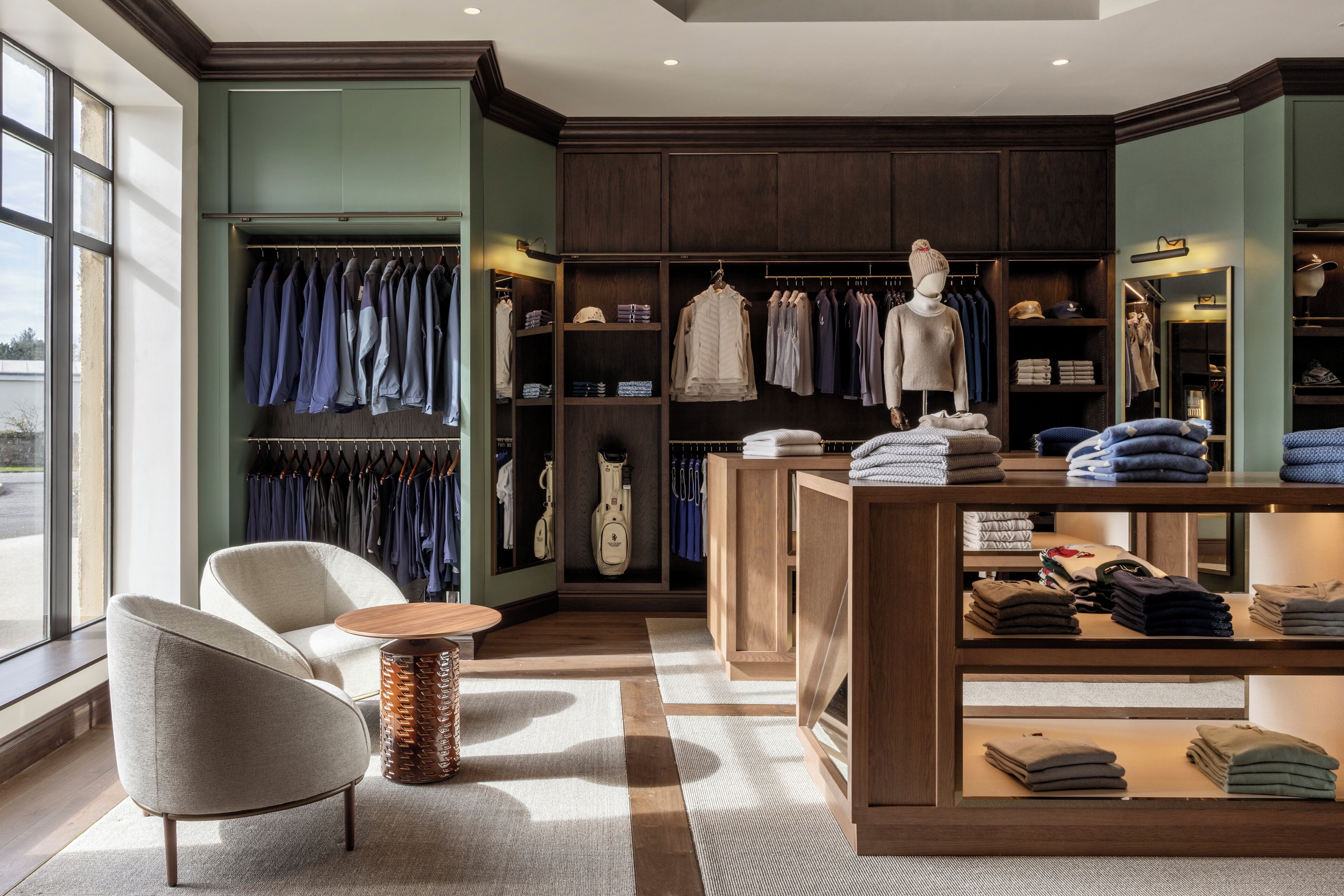 A well-lit boutique hotel room with a large window, a seating area with a chair and a small round table, and a wardrobe with neatly folded clothes and a mannequin.