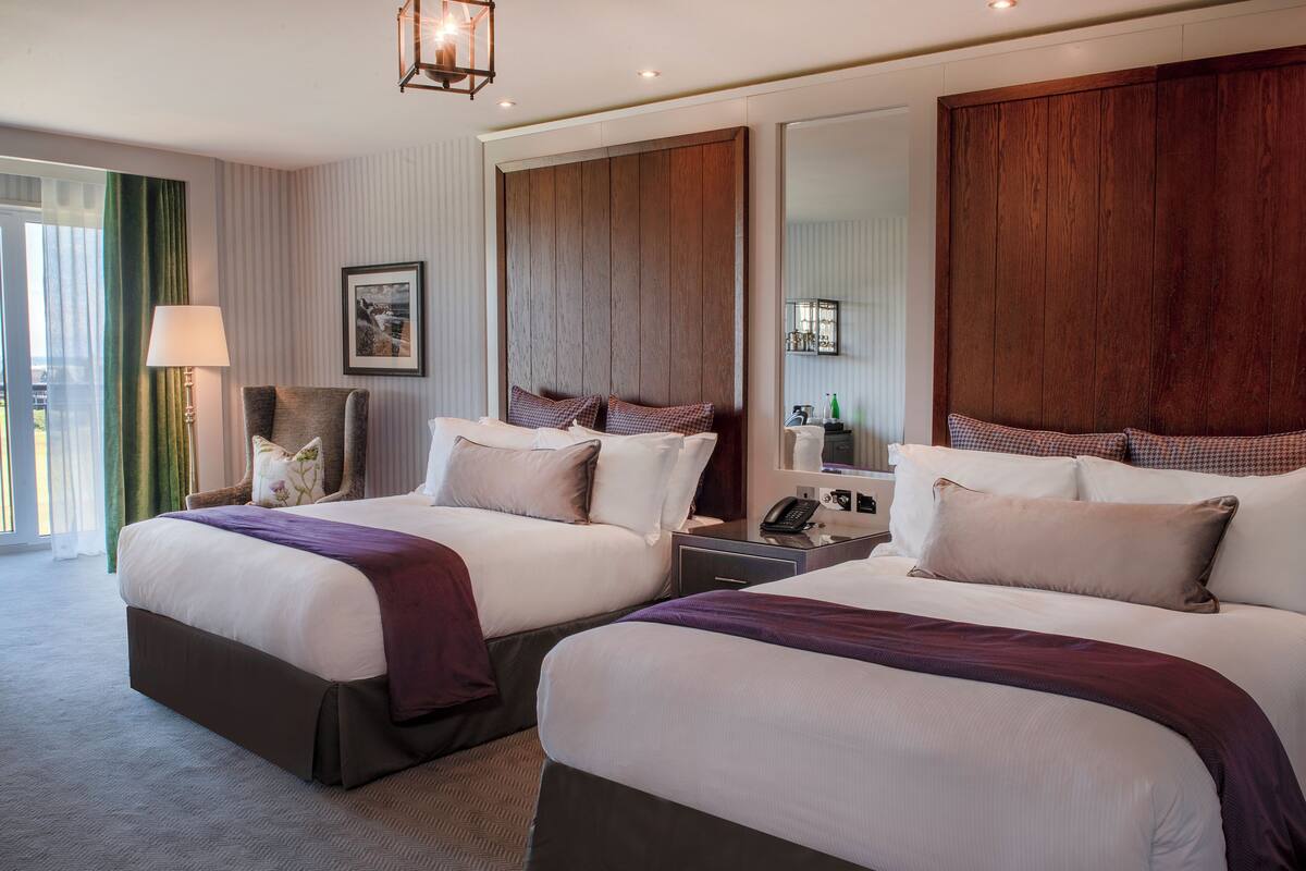 A hotel room with two beds, a large mirror, a nightstand, a lamp, and a view of the outdoors.