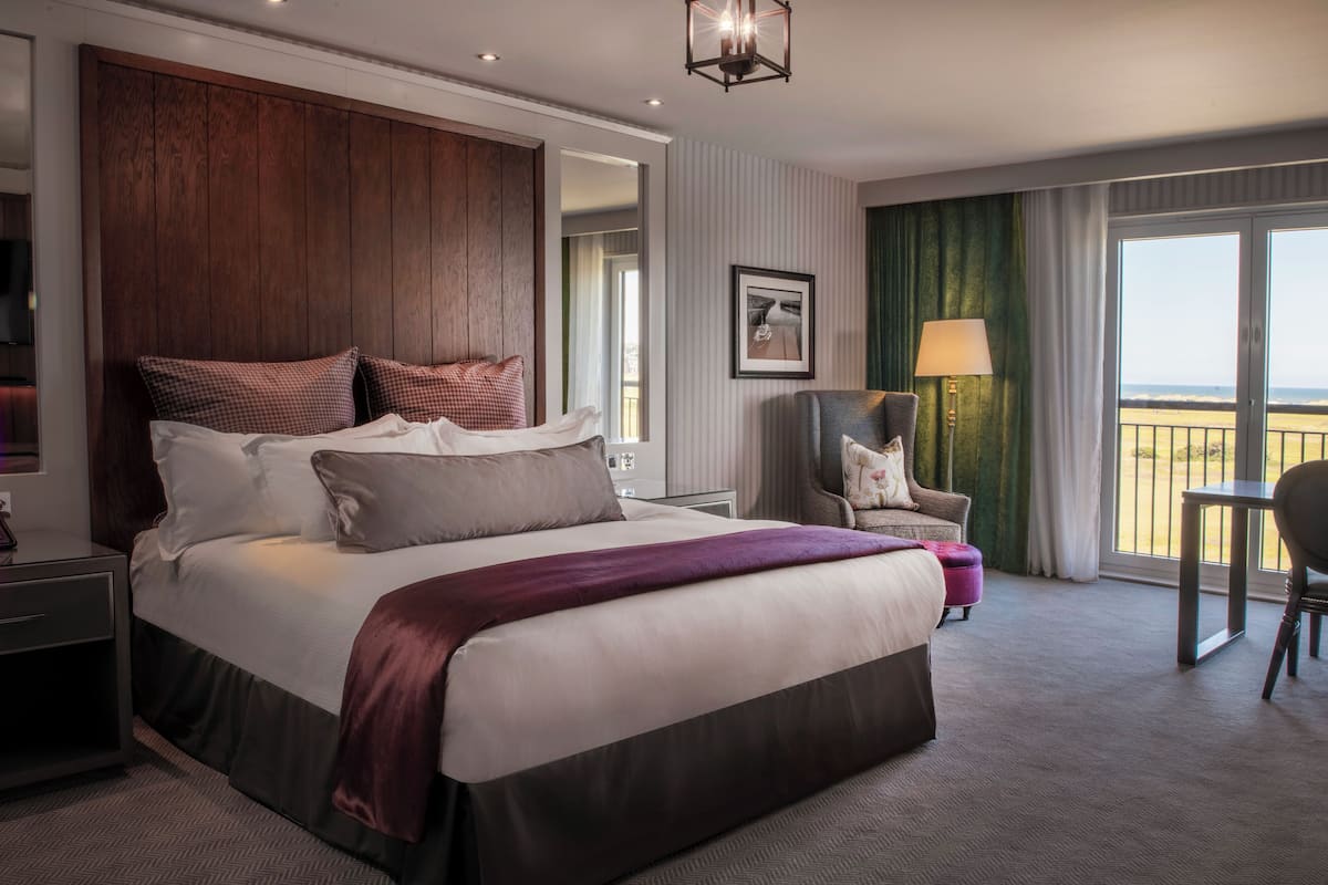 A hotel room with a large bed, a chair, a desk, and a balcony with a view.