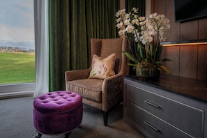 A room with a view of a grassy field, a brown armchair with a floral pillow, a purple ottoman, and a wooden cabinet with a TV mounted above it.