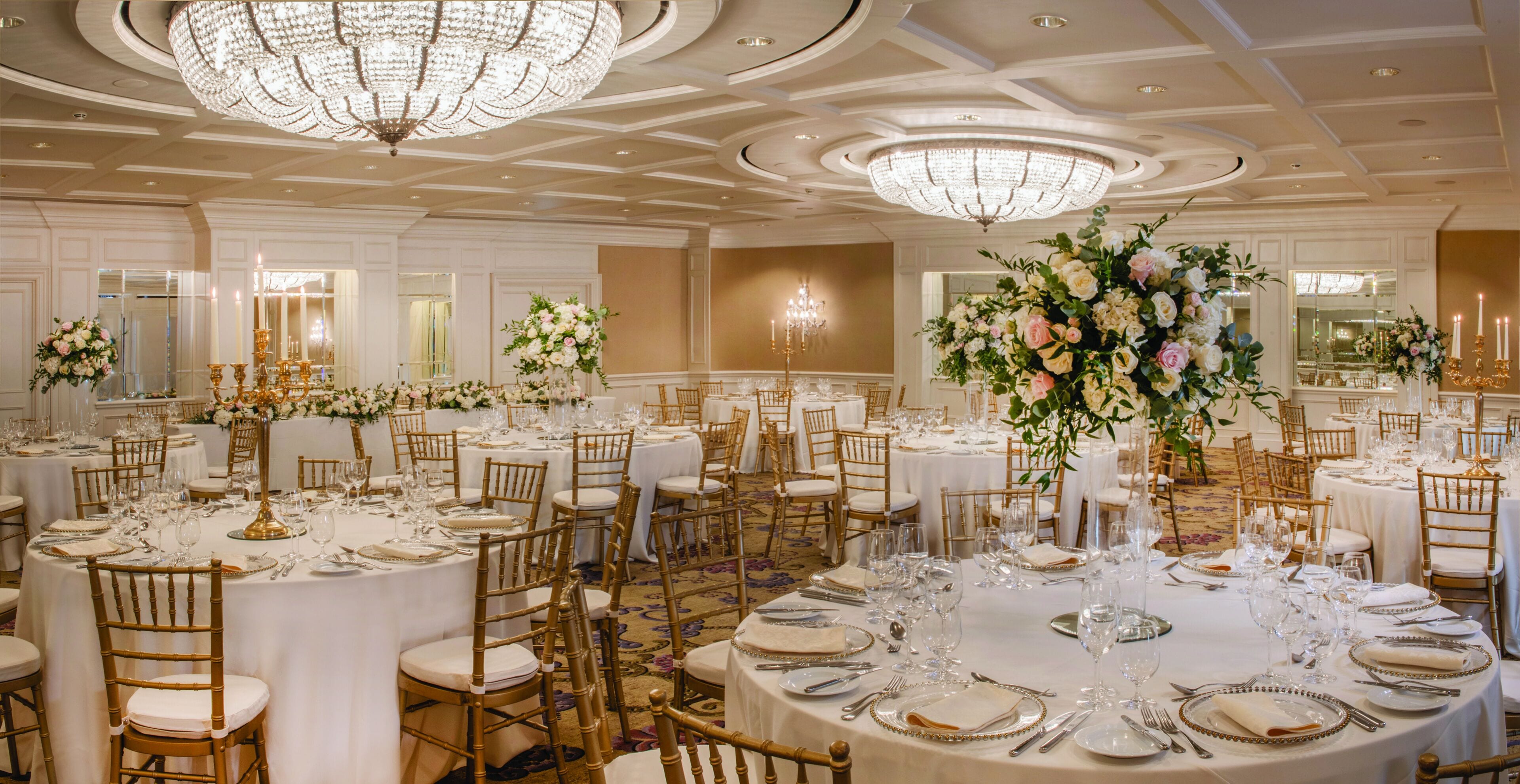 Ein Bankettsaal mit runden Tischen, festlich für eine Veranstaltung gedeckt, mit goldenen Stühlen, weißen Tischdecken und floralen Mittelpunkten.