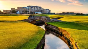 A golf course with a stone bridge over a water channel, a large building in the background, and a sunset sky. - Old Course Hotel (St. Andrews)
