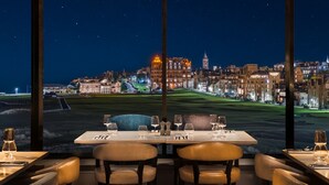 A dining area with a table set for two, large windows offering a nighttime view of a cityscape with illuminated buildings and a golf course. - Old Course Hotel (St. Andrews)