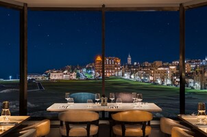 Un área de comedor con una mesa preparada para dos, ventanas amplias que ofrecen una vista nocturna de un paisaje urbano con edificios iluminados y un campo de golf.