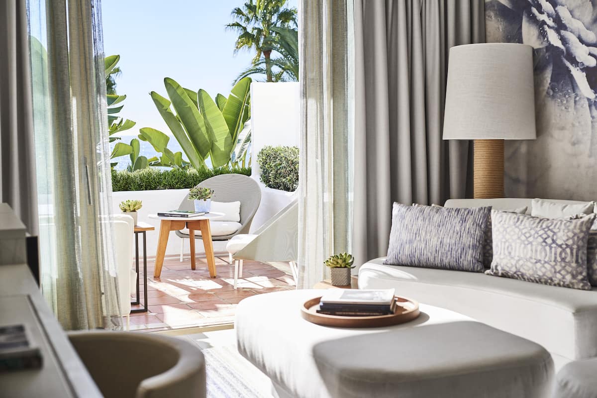 A modern hotel room with a large bed, a sitting area with a chair and table, a lamp, and a view of the outdoors.