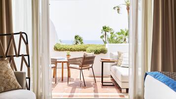 Eine gemütliche Terrasse mit einer weißen Couch, einem Korbstuhl und einem kleinen Tisch. Durch die Vorhänge sind eine Holzpergola und der Blick aufs Meer zu sehen.