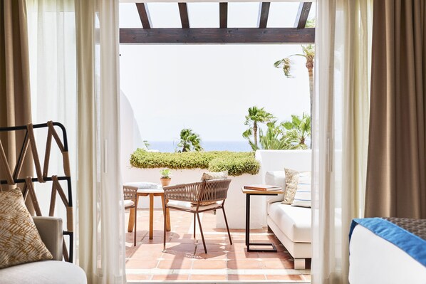 A cozy patio with a white sofa, a wicker chair, and a small table. A wooden pergola and a view of the sea are visible through the curtains. - Puente Romano Marbella (Marbella)