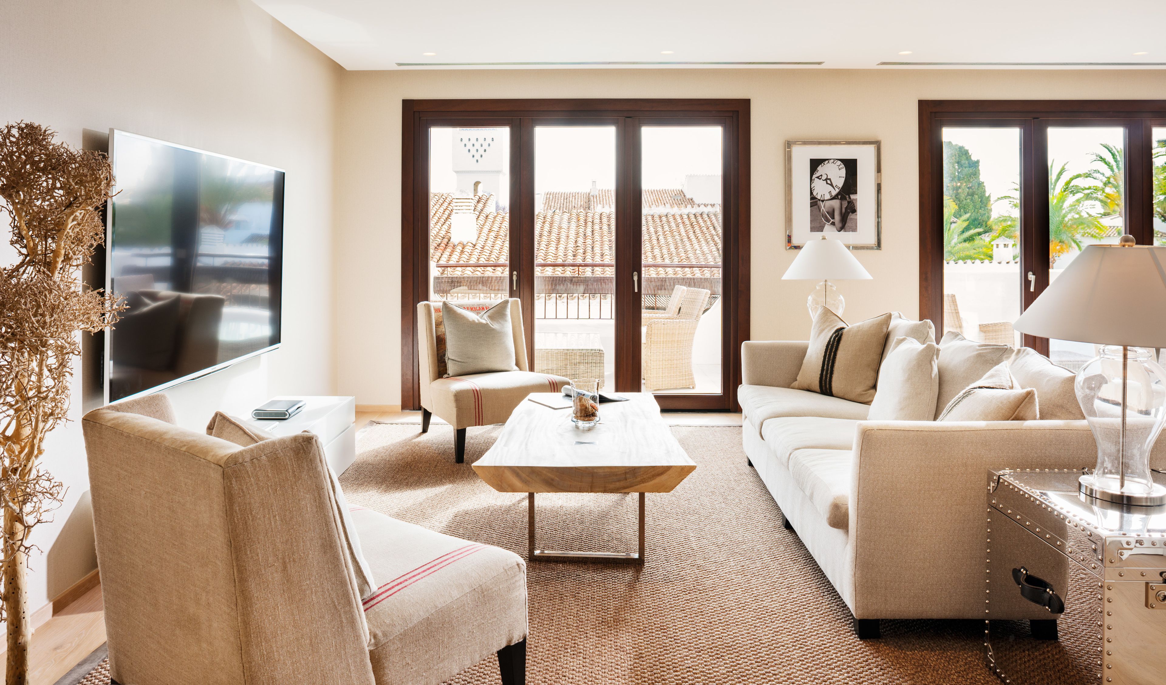 Un salón moderno con un televisor de pantalla plana, dos sofás, una mesa de centro y un amplio ventanal con vistas a un edificio de tejado a ladrillo.