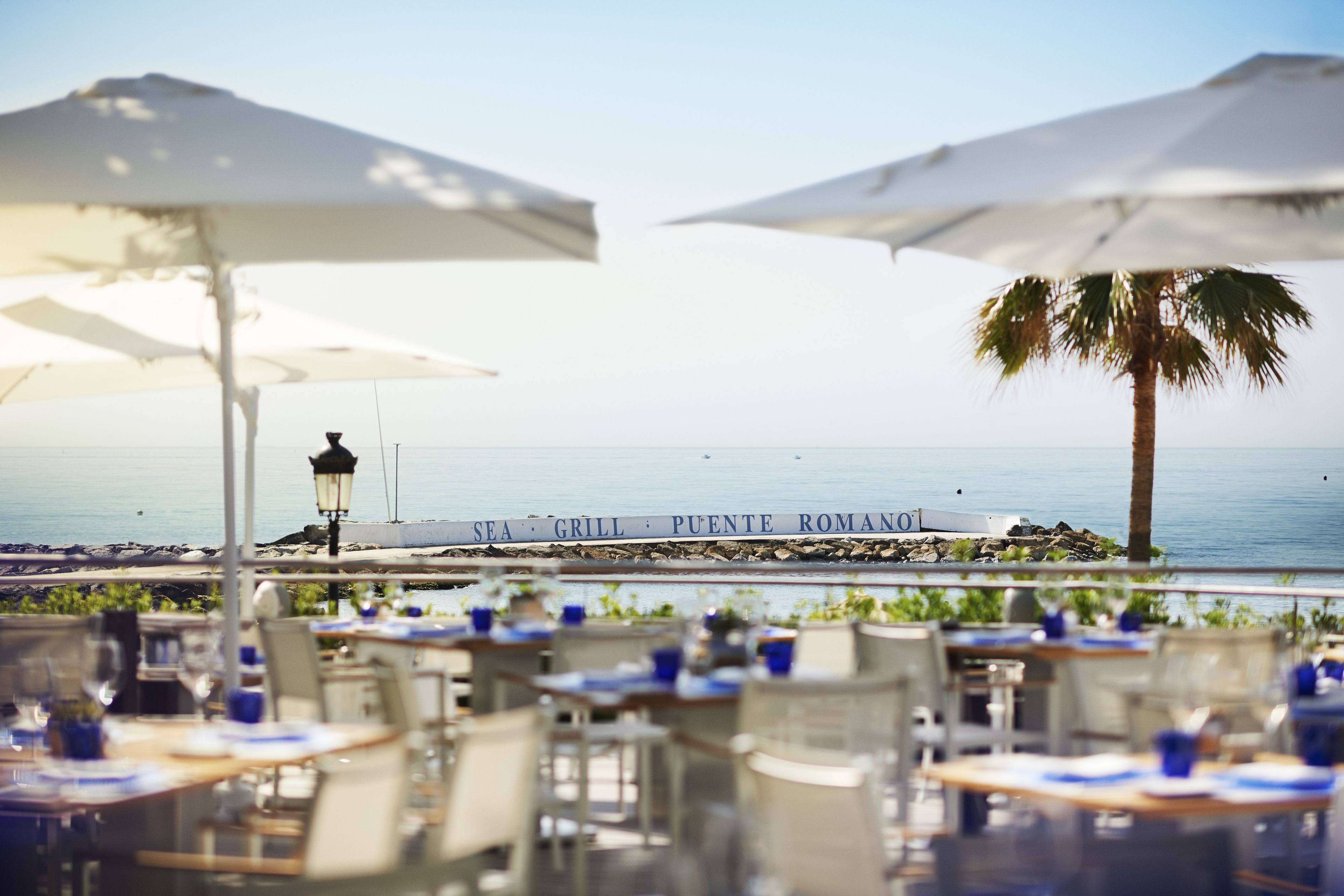 Un área de comedor al aire libre con mesas y sombrillas, con vista a una playa. Se observa un letrero que dice "SEA GRILL PUENTE ROMANO".