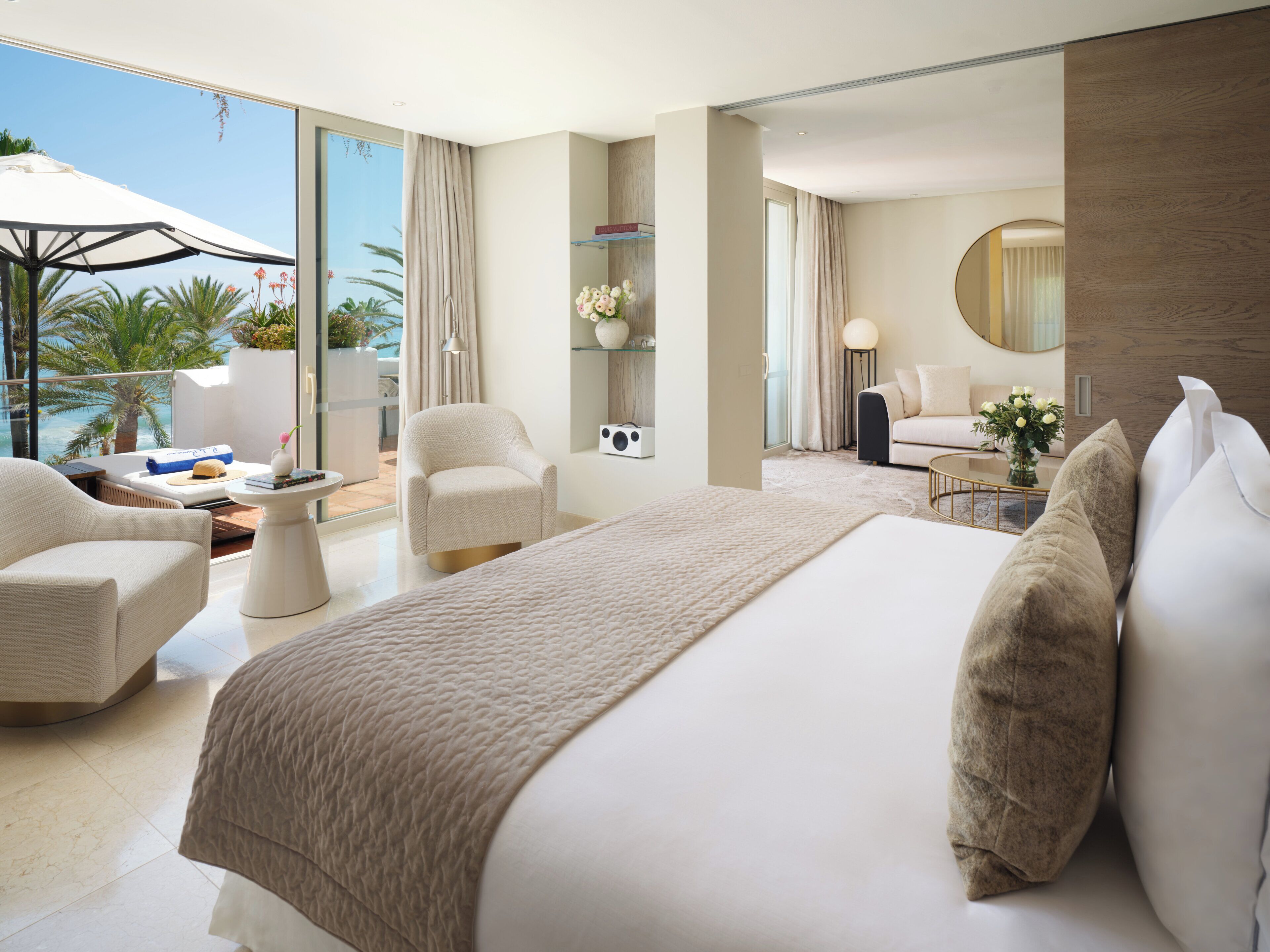 Ein modernes Hotelzimmer mit einem großen Bett, einer Sitzecke mit Sofa und Sesseln, einem kleinen Tisch und Blick ins Freie.
