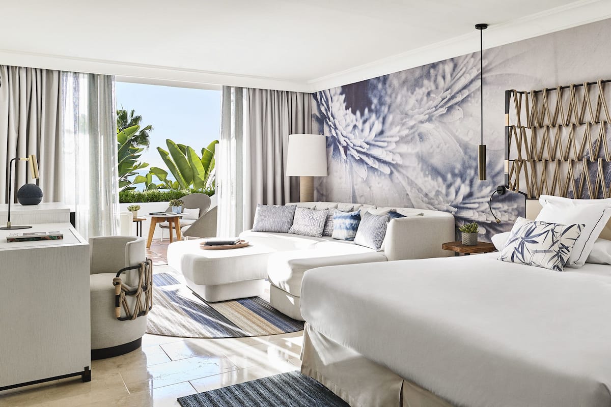 A hotel room with two beds, a sofa, a desk, and a large window with curtains.