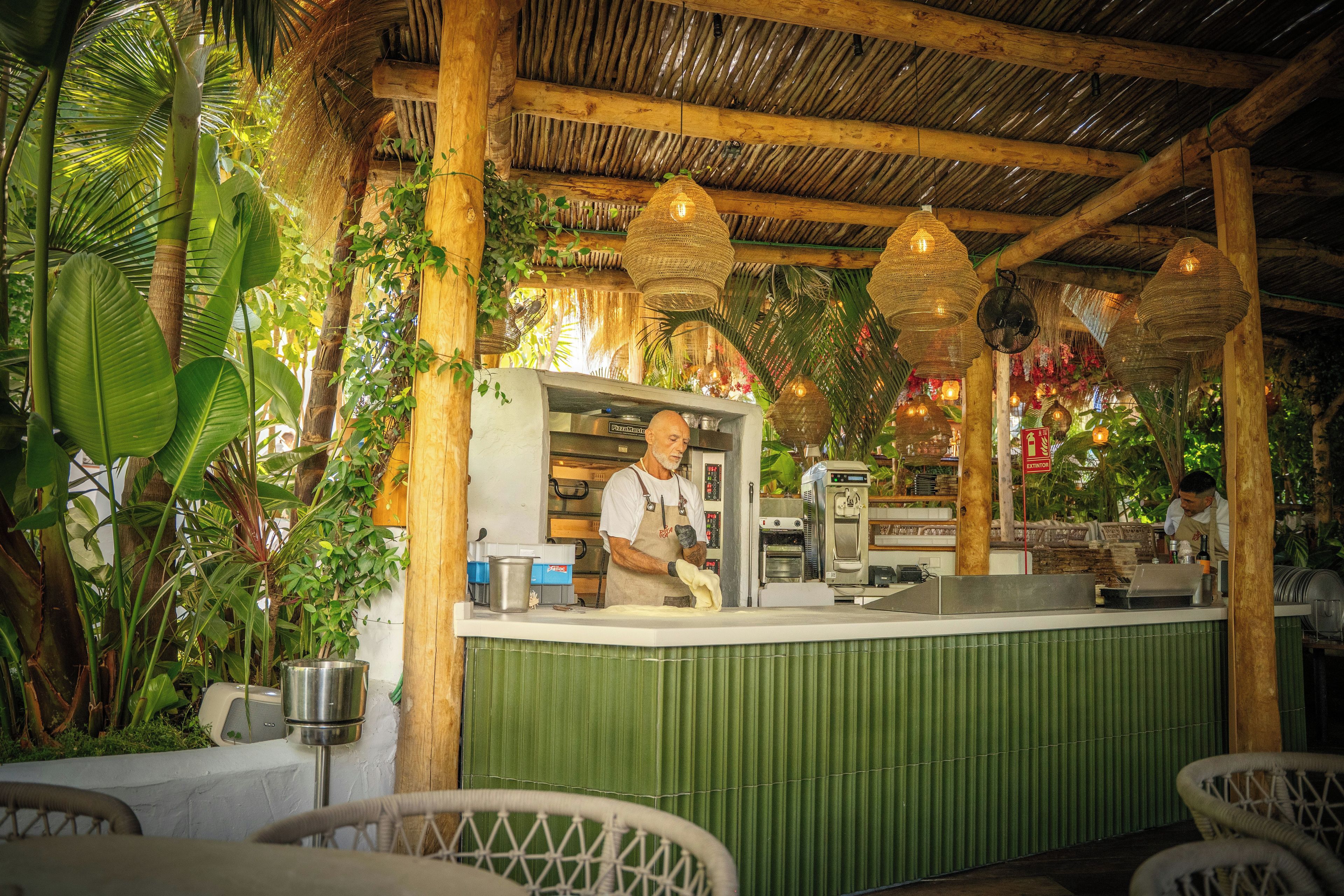 En utomhusrestaurang med en träkonstruktion, en bardisk där kocken arbetar och diverse köksutrustning.