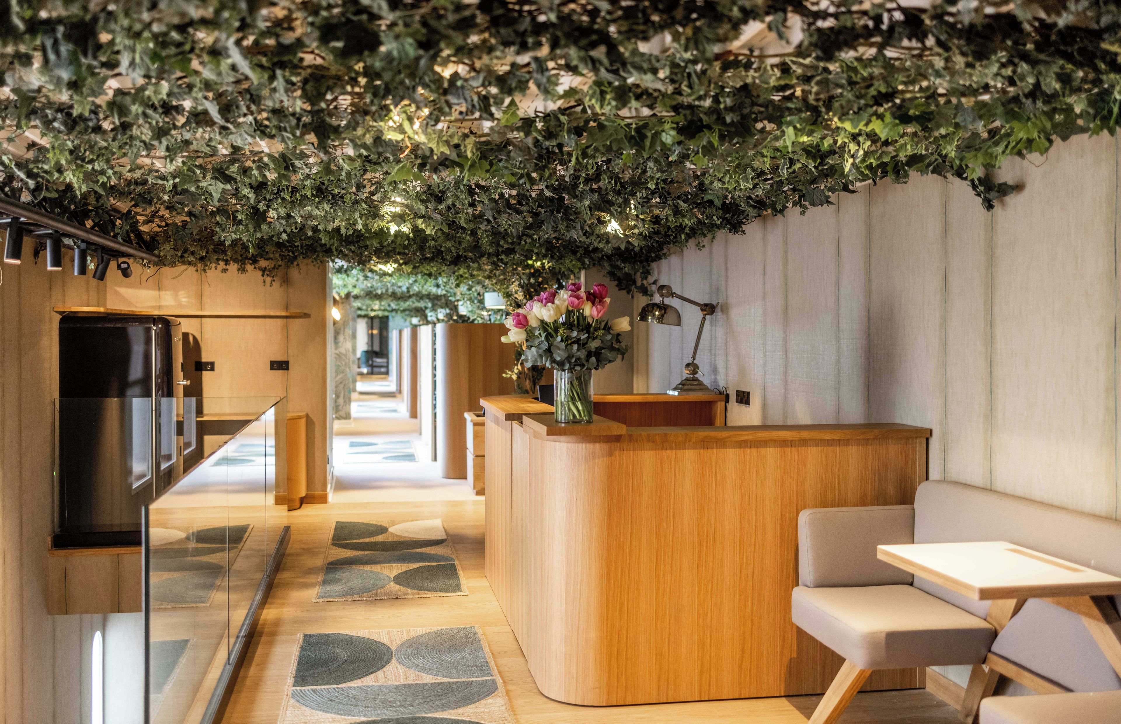 A reception area with a wooden reception desk, a vase with flowers, a desk lamp, and a seating area with a sofa and a chair.
