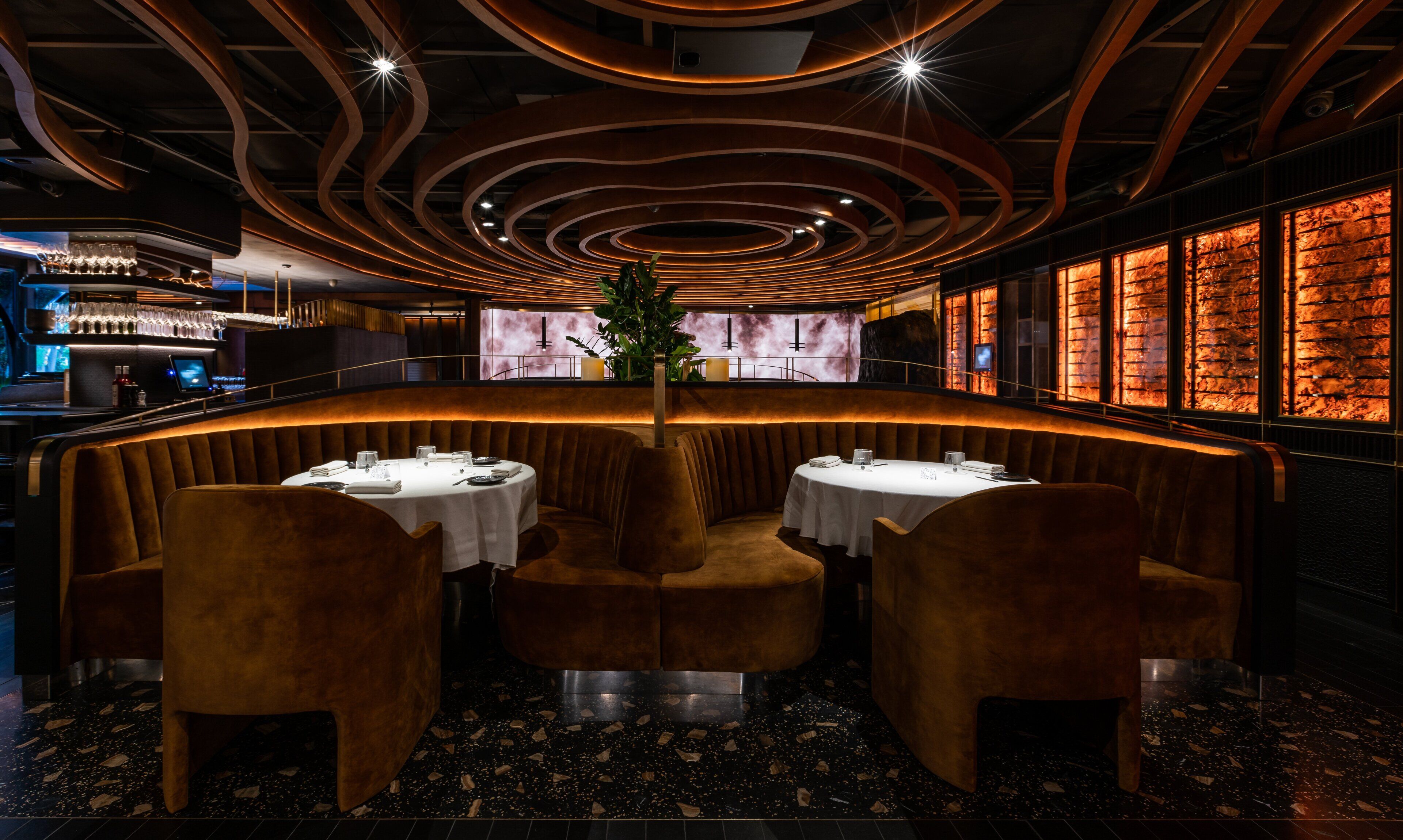 Un restaurante con una decoración moderna, que presenta una zona de estar curvada con cabinas de terciopelo y una mesa central.