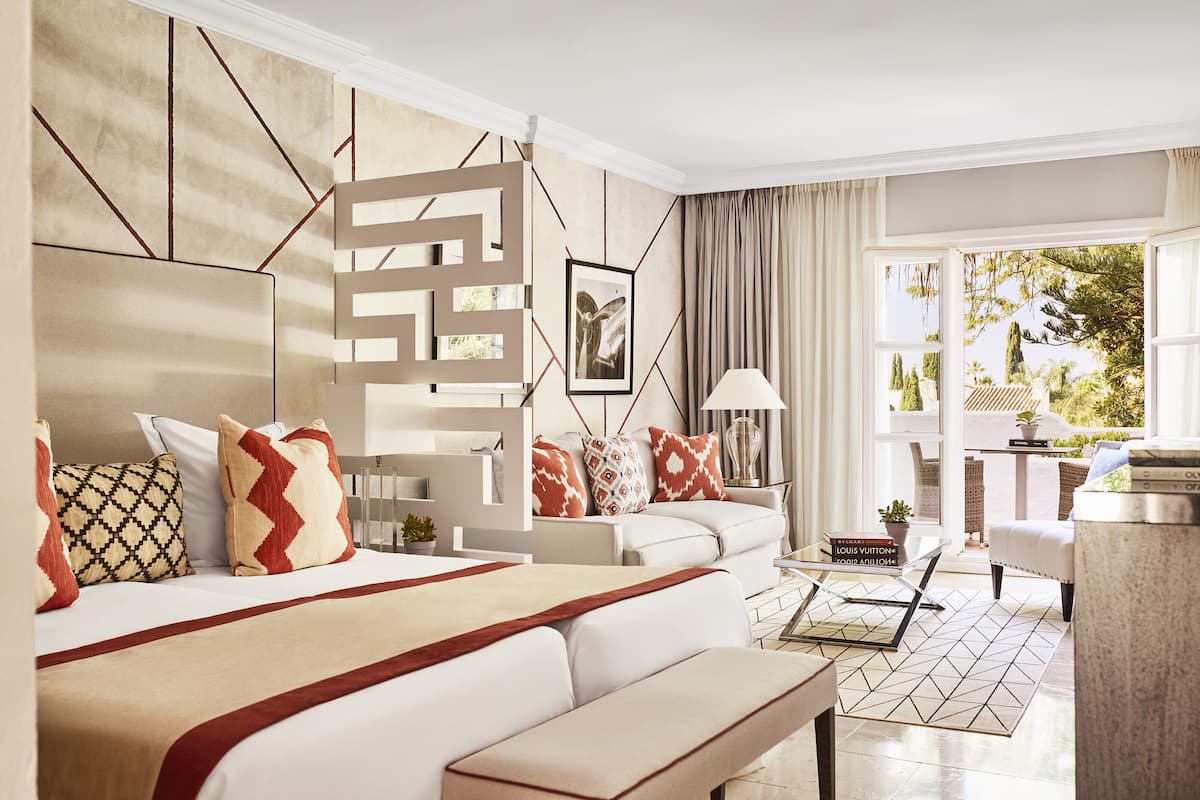 A modern hotel room with a large bed, a sofa, a desk, and a view of the outdoors.