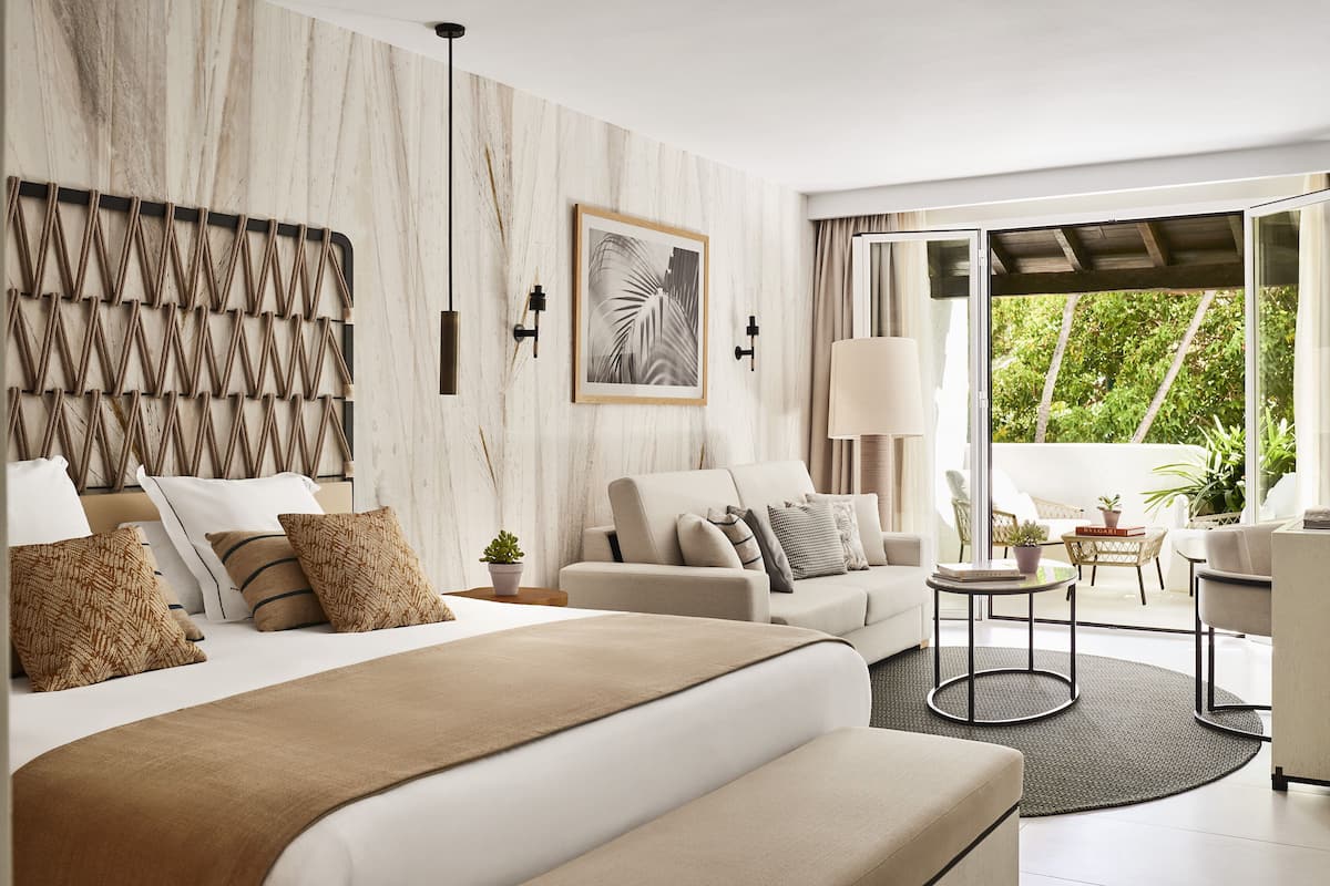 A modern hotel room with a large bed, a sofa, a small table, and a view of the outdoors.