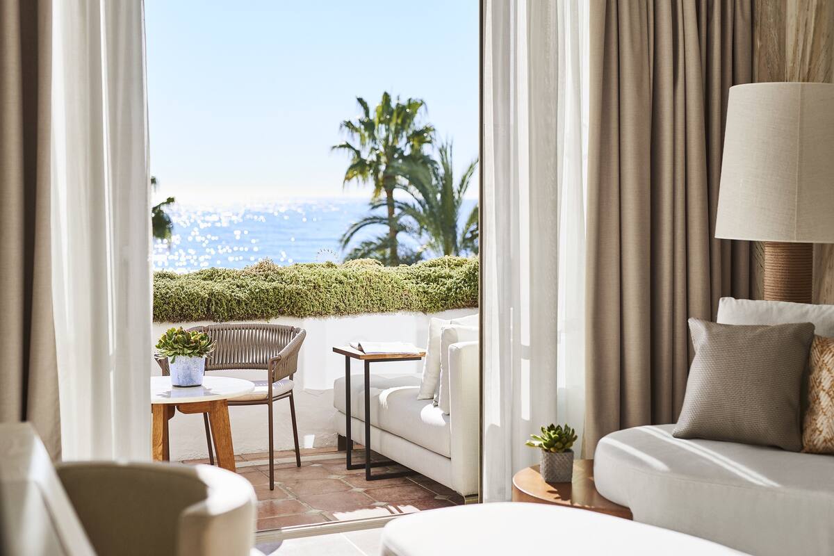 A hotel room with a balcony, a sofa, a chair, a small table, and a lamp.