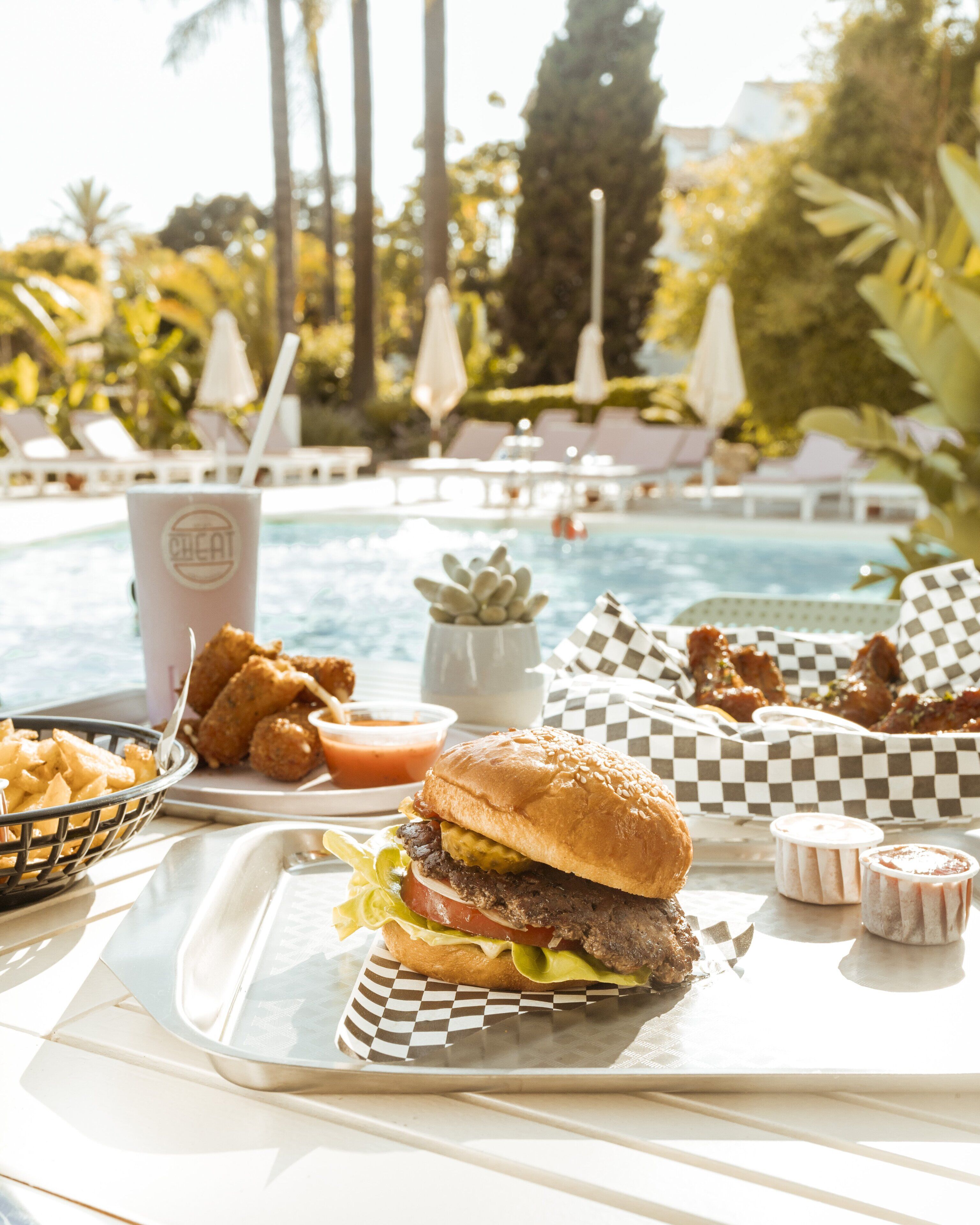 Un plateau repas comprenant un hamburger, des frites et des boissons, placé près d’une piscine, avec des chaises longues et des parasols en arrière-plan.