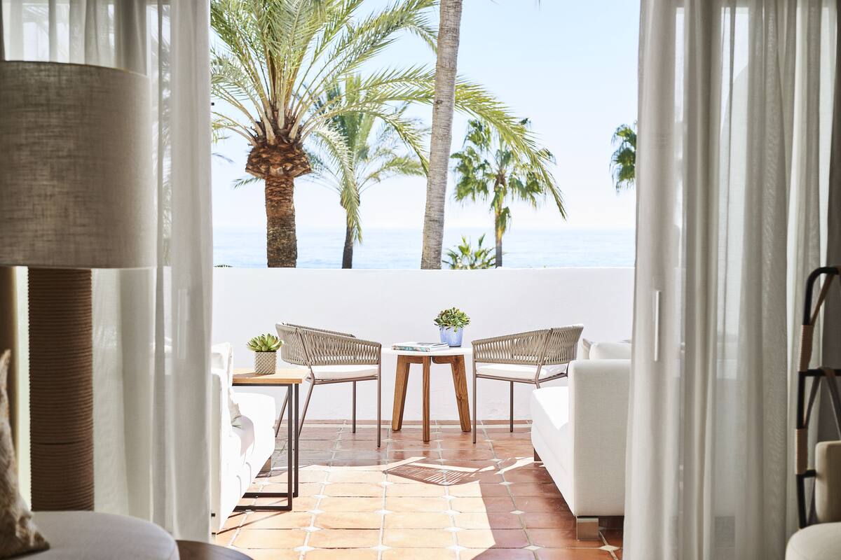 A balcony with a white sofa, two wicker chairs, a small table with a plant, and a view of palm trees and the sea.