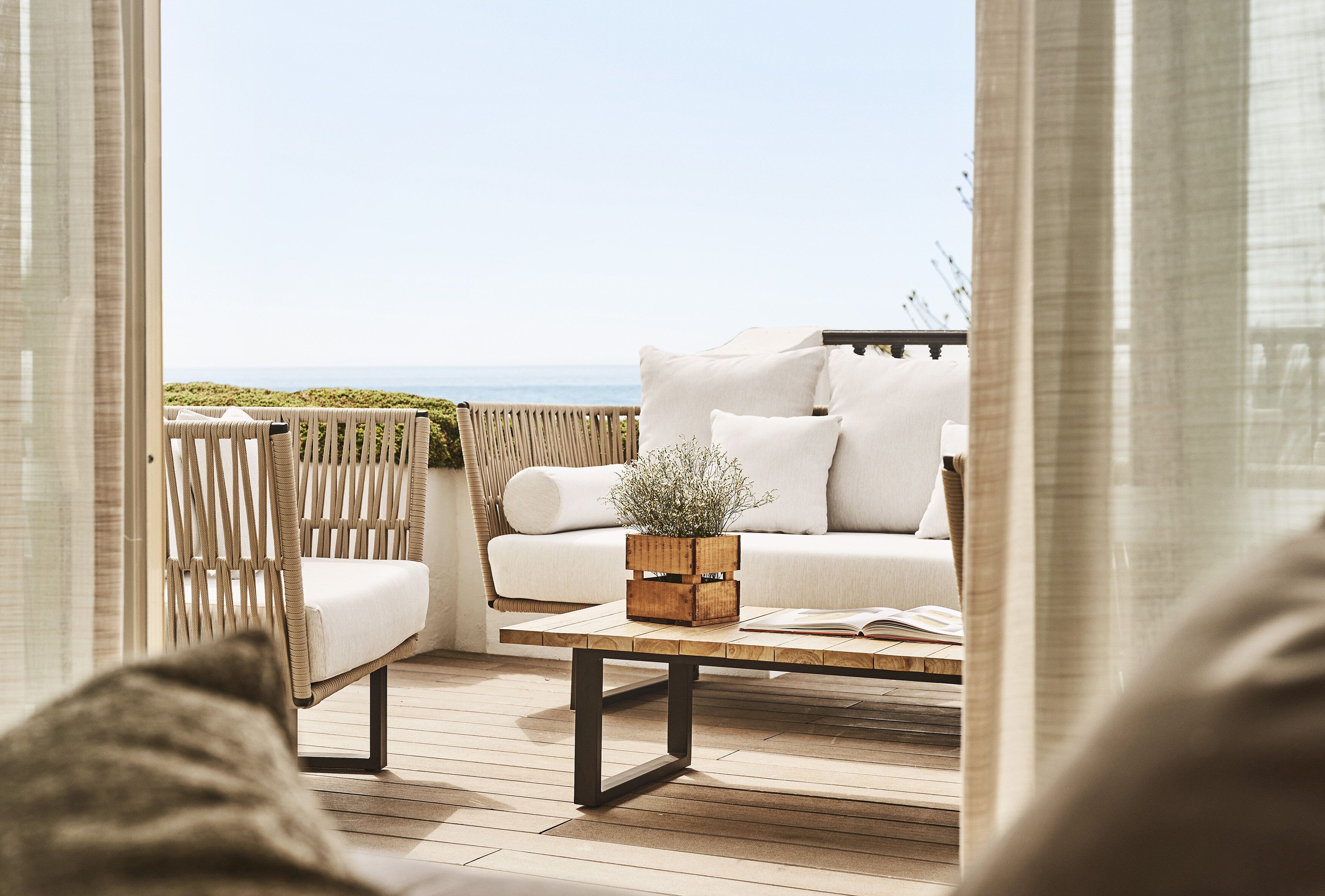 Een houten terras met een rieten stoel, een witte bank en een houten salontafel met een plant.