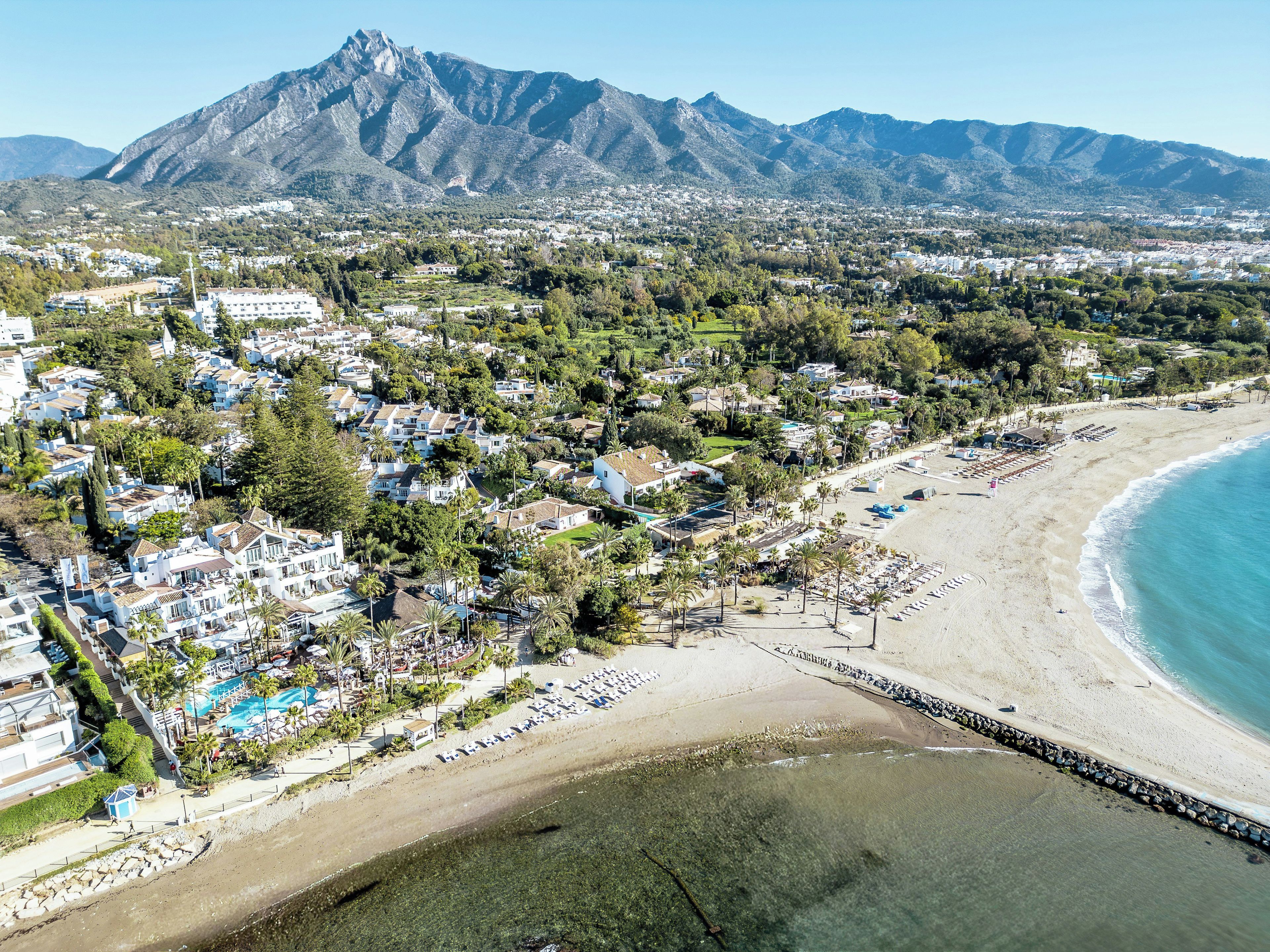 Un pueblo costero con playa, edificios residenciales y montañas al fondo.