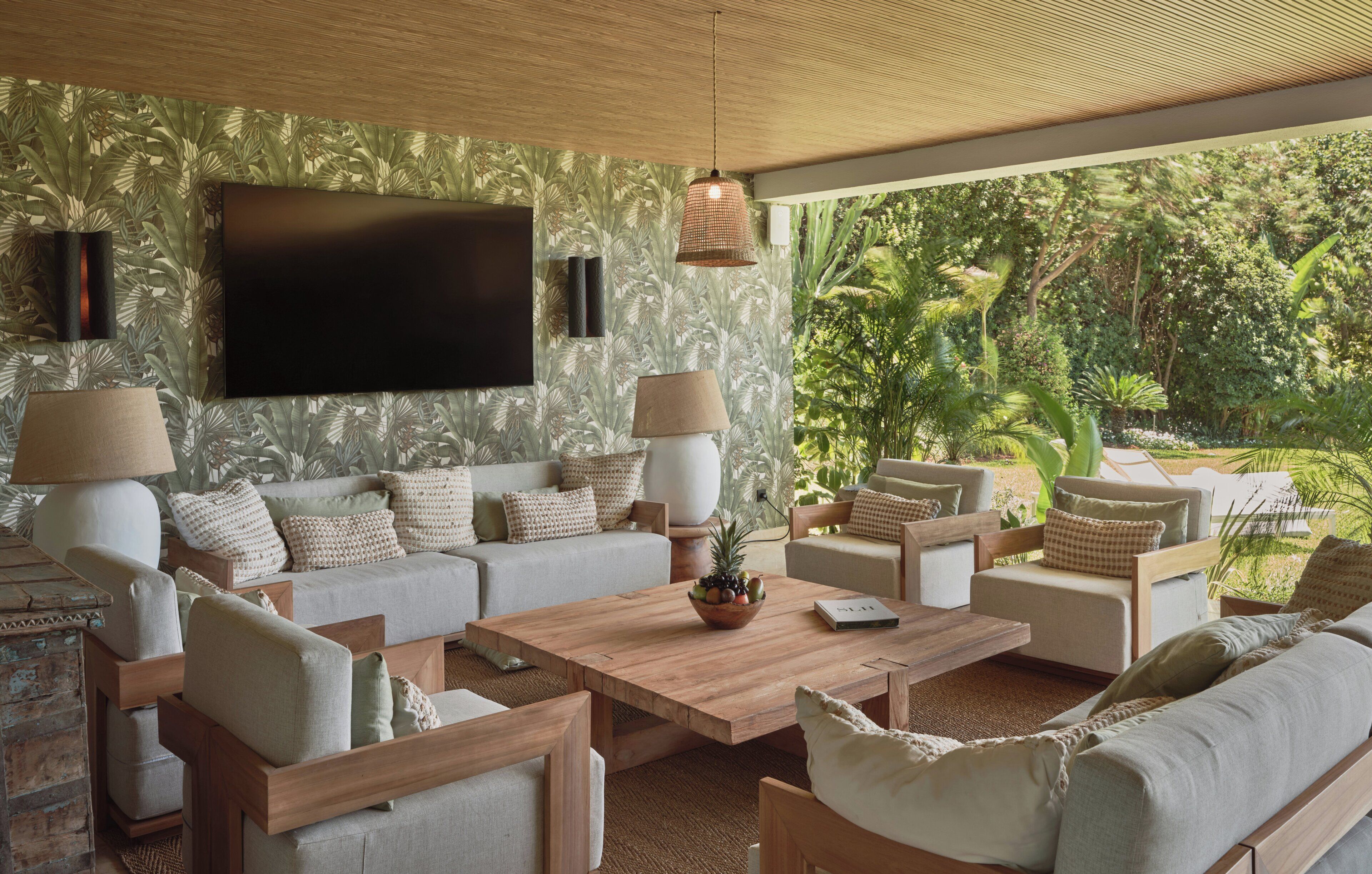 A living room with a large flat-screen TV, two floor lamps, a wooden coffee table, and a view of lush greenery outside.