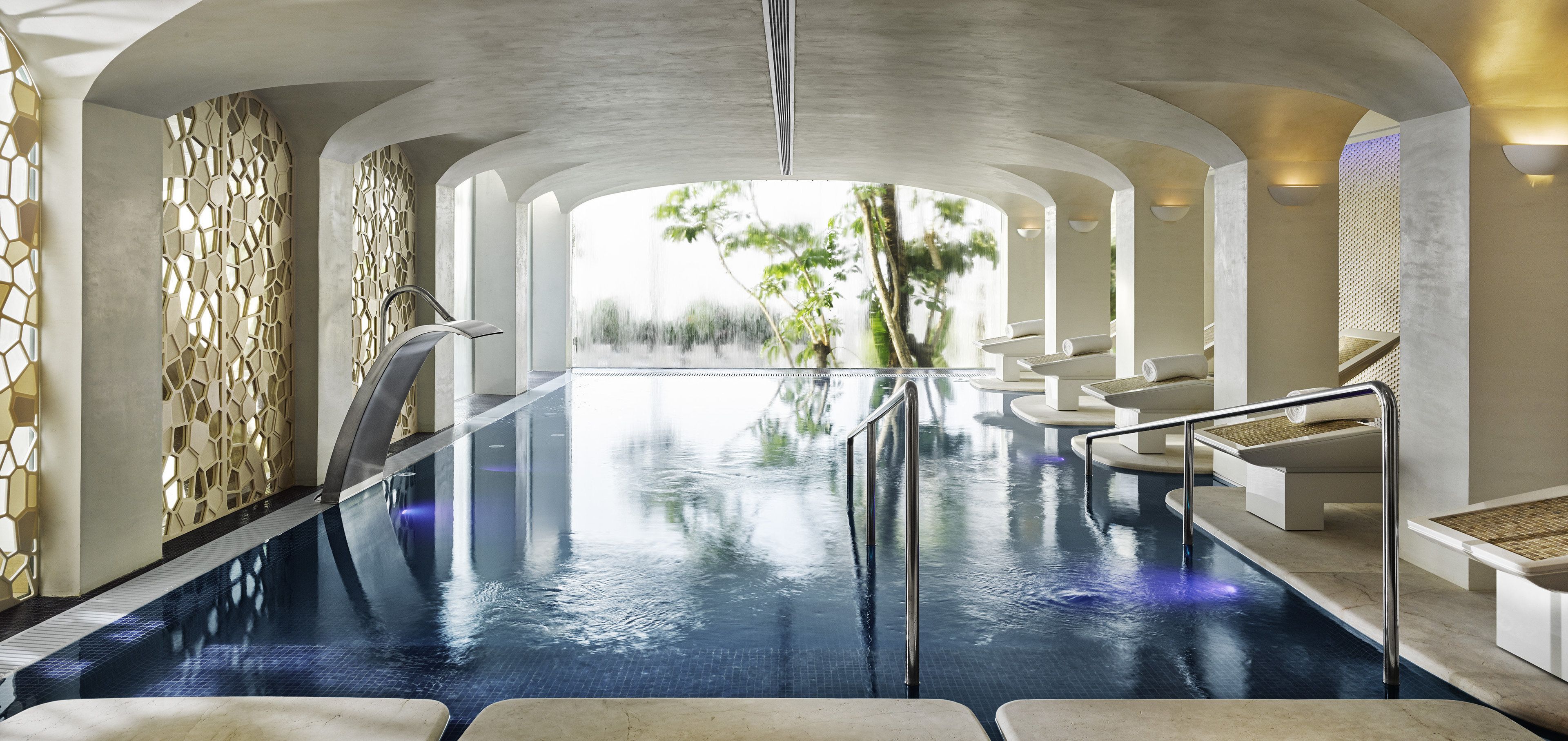 Piscina infinita en el interior con vista a árboles, rodeada de paredes blancas y zonas de estar.
