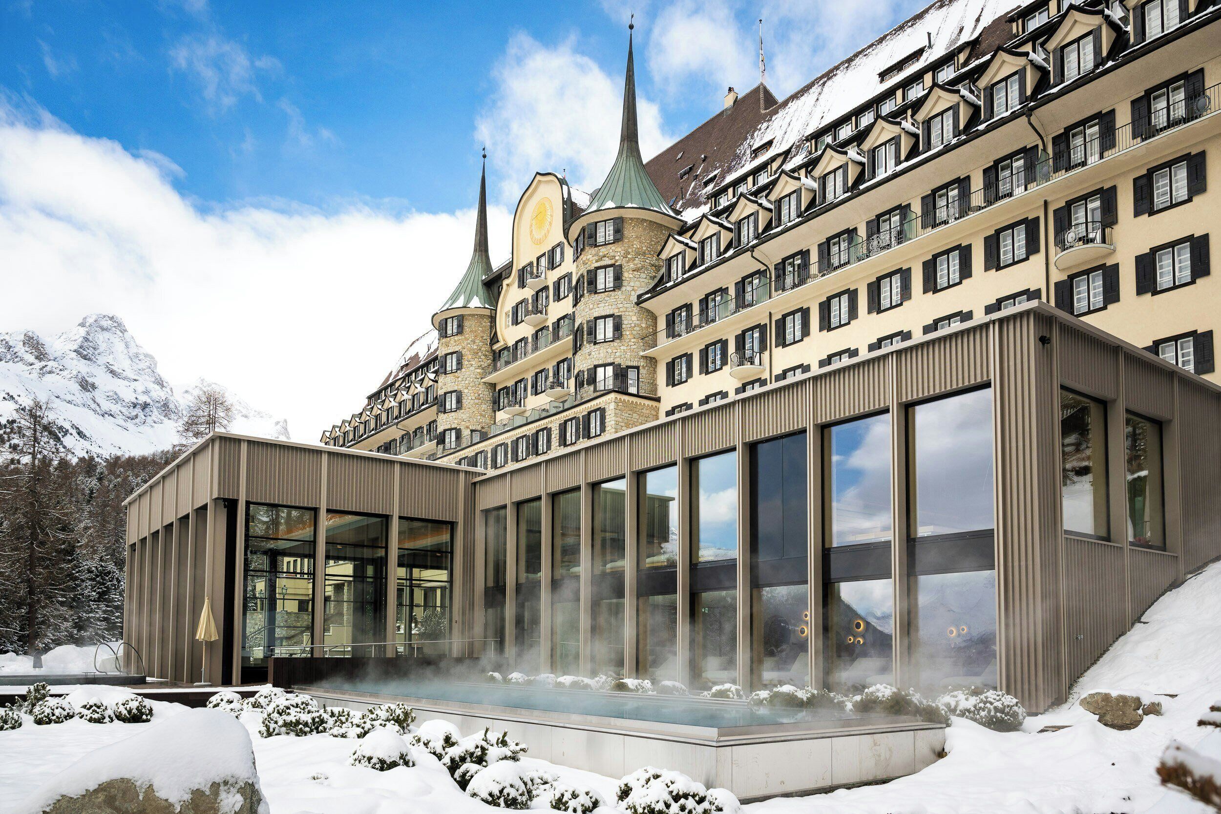 A modern hotel with a glass facade, a traditional building with spires in the background, and snow-covered surroundings.