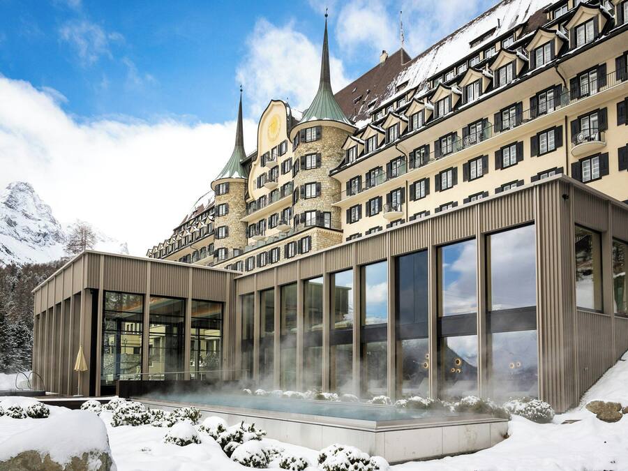 A modern hotel with a glass facade, a traditional building with spires in the background, and snow-covered surroundings.
