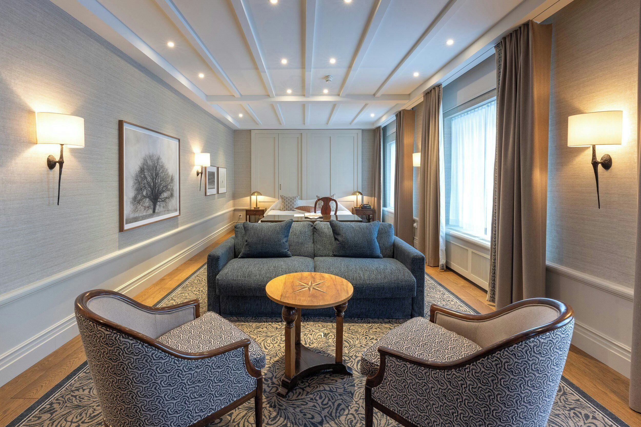 A hotel room with a blue sofa, two armchairs, a small round table, and wall-mounted lamps.