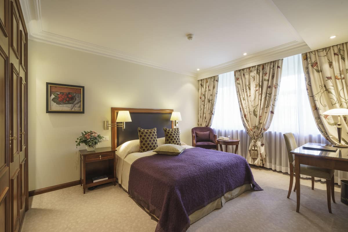 A hotel room with a large bed, two bedside tables with lamps, a chair, a desk, and a window with curtains.