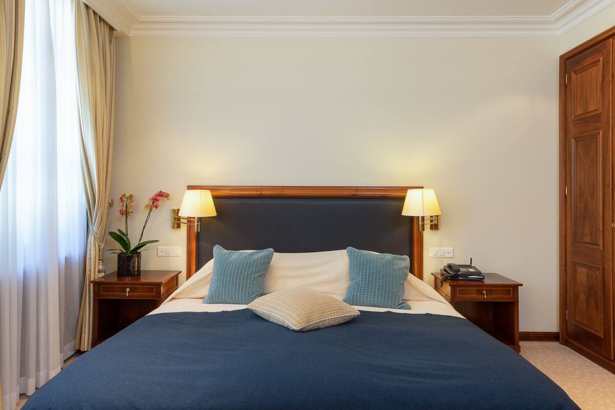 A neatly made bed with a dark blue bedspread, two pillows, and a bedside table with a lamp.