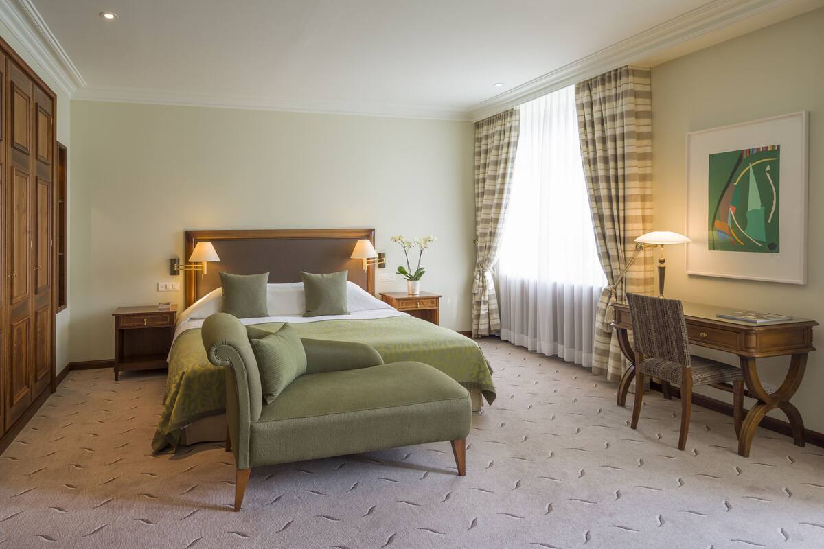 A hotel room with a large bed, a desk, a chair, a nightstand, and a window with curtains.