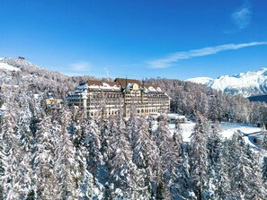 A large, multi-story hotel with a traditional European architectural style, surrounded by snow-covered trees and mountains in the background. - Suvretta House (St. Moritz)