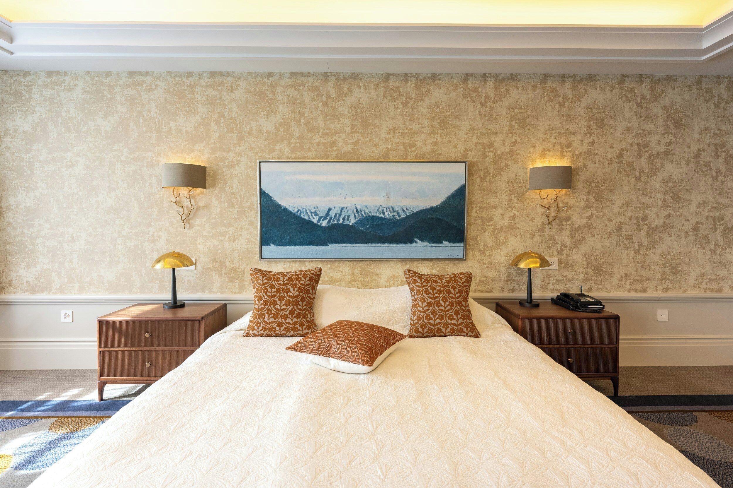 Una cama bien hecha con almohadones estampados, dos lámparas de noche y un cuadro enmarcado con montañas en la pared.