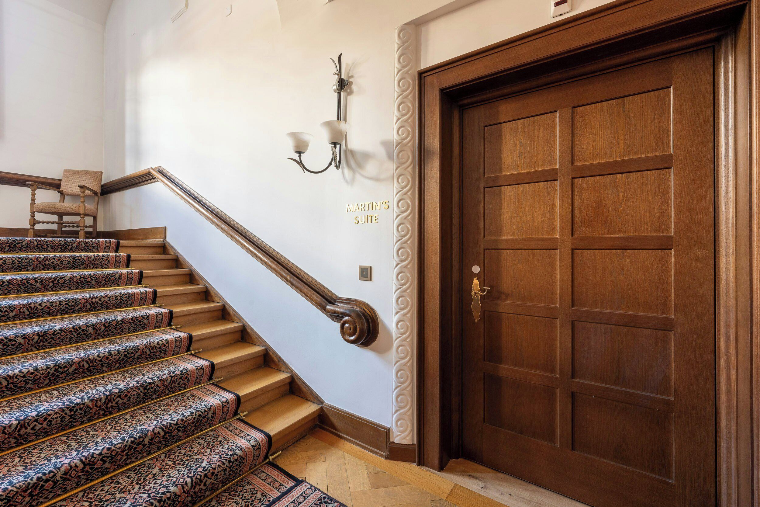 Un escalier en bois, recouvert d’une moquette à motifs, mène à une porte sur laquelle on peut lire : « Suite de Martin ».
